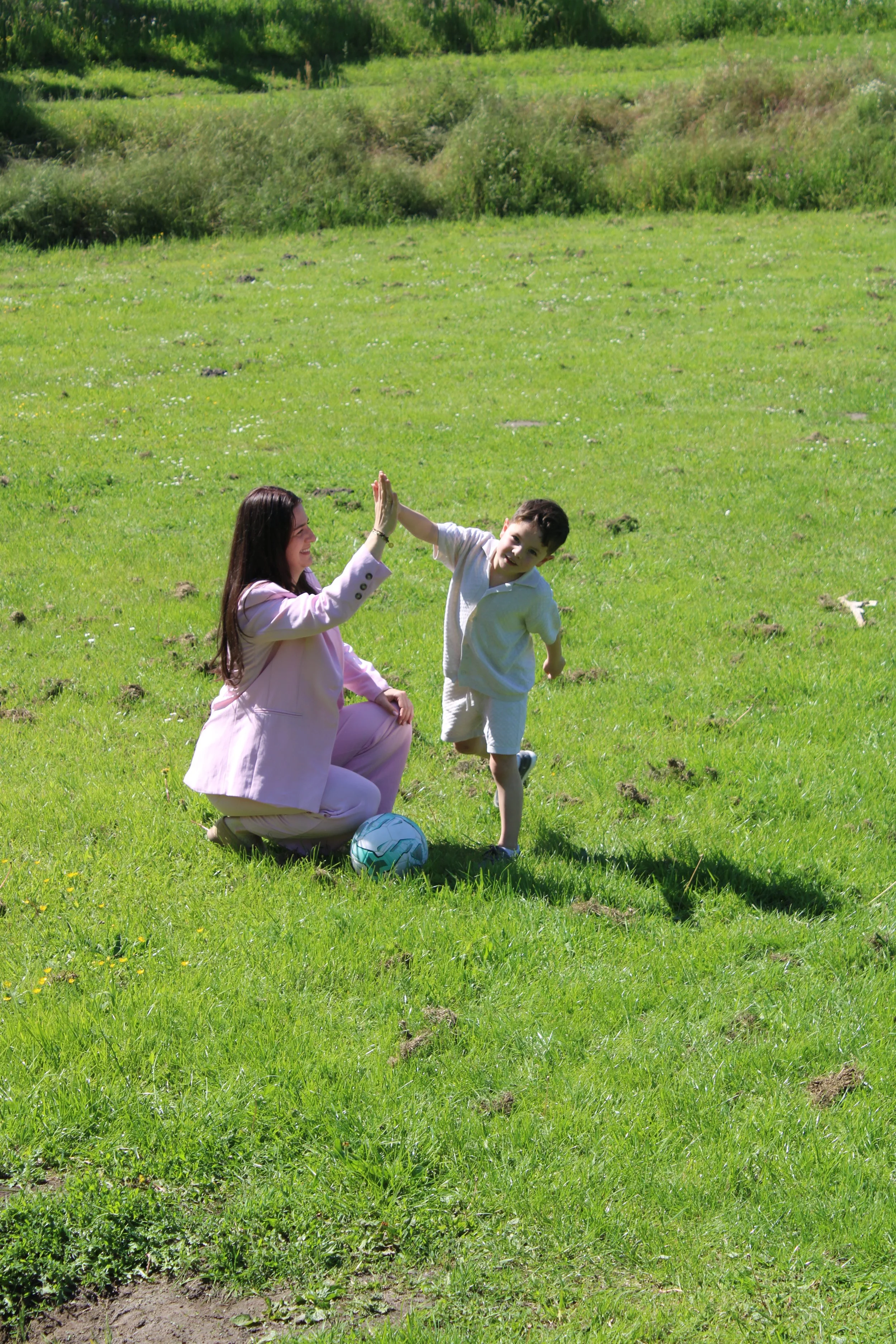 A woman kneeling on green grass and a young boy playing soccer on a sunny day in a park with trees and bushes in the background.