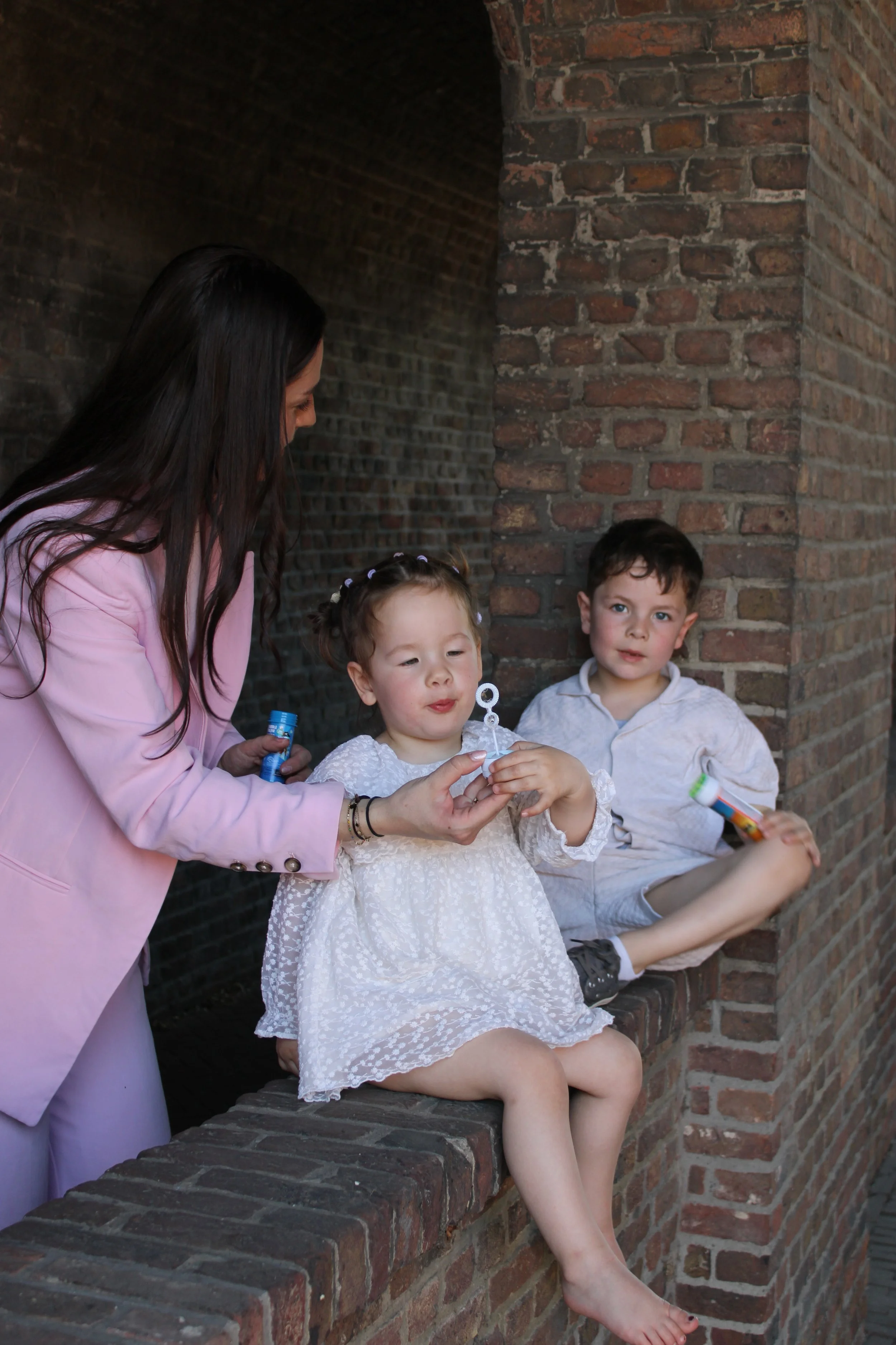 A woman in a pink blazer talking to two children sitting on a brick ledge under a tunnel. The girl has curly hair in pigtails, wearing a white dress, and the boy has short hair, wearing a gray hoodie. The girl is holding a bubble wand, and the boy is holding a bubble solution bottle. The girl is sitting with her legs crossed, and the boy's foot is resting on the brick ledge.