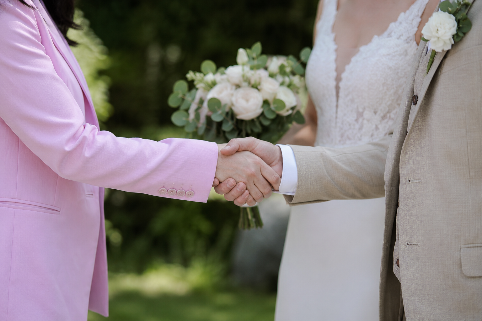Close-up of a same-sex wedding handshake, with one person in a pink suit and the other in a beige suit, holding a wedding bouquet in the background.