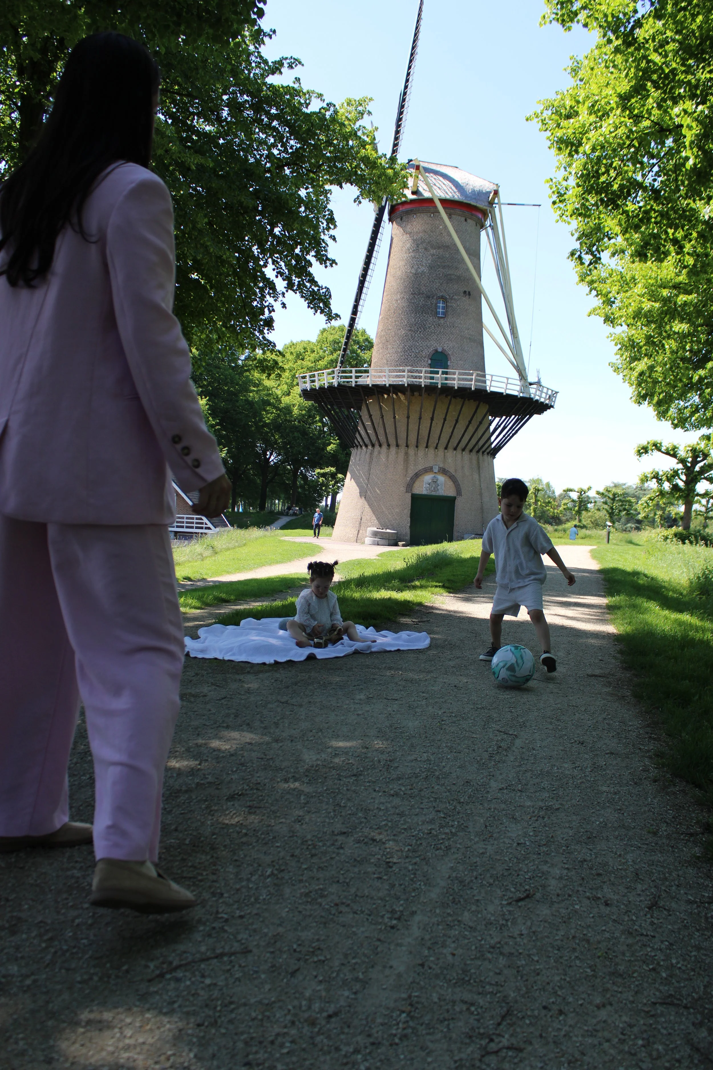 Children playing soccer outdoors in a park near a windmill, with trees and a clear blue sky in the background.