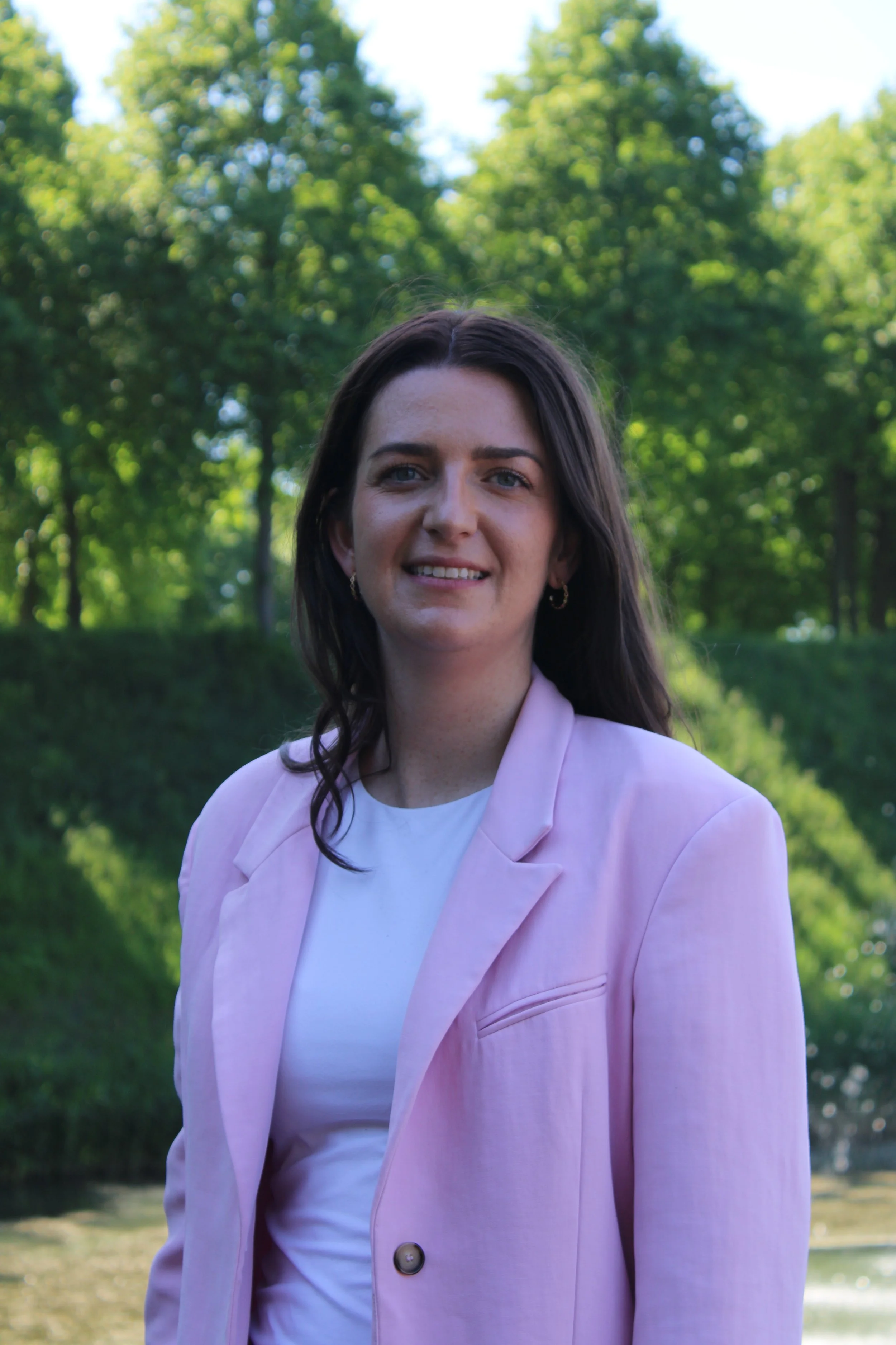 A woman with long dark hair and blue eyes is smiling outdoors with green trees and a river in the background. She is wearing a light pink blazer over a white top.