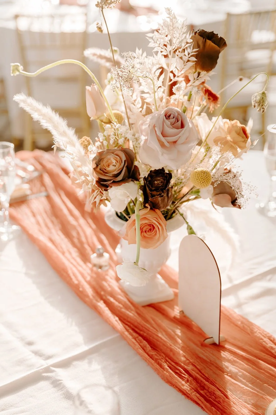 Elegant flower centerpiece with various roses, dried flowers, and pampas grass in a white vase on a table with a peach-colored table runner, set in a well-lit event space.
