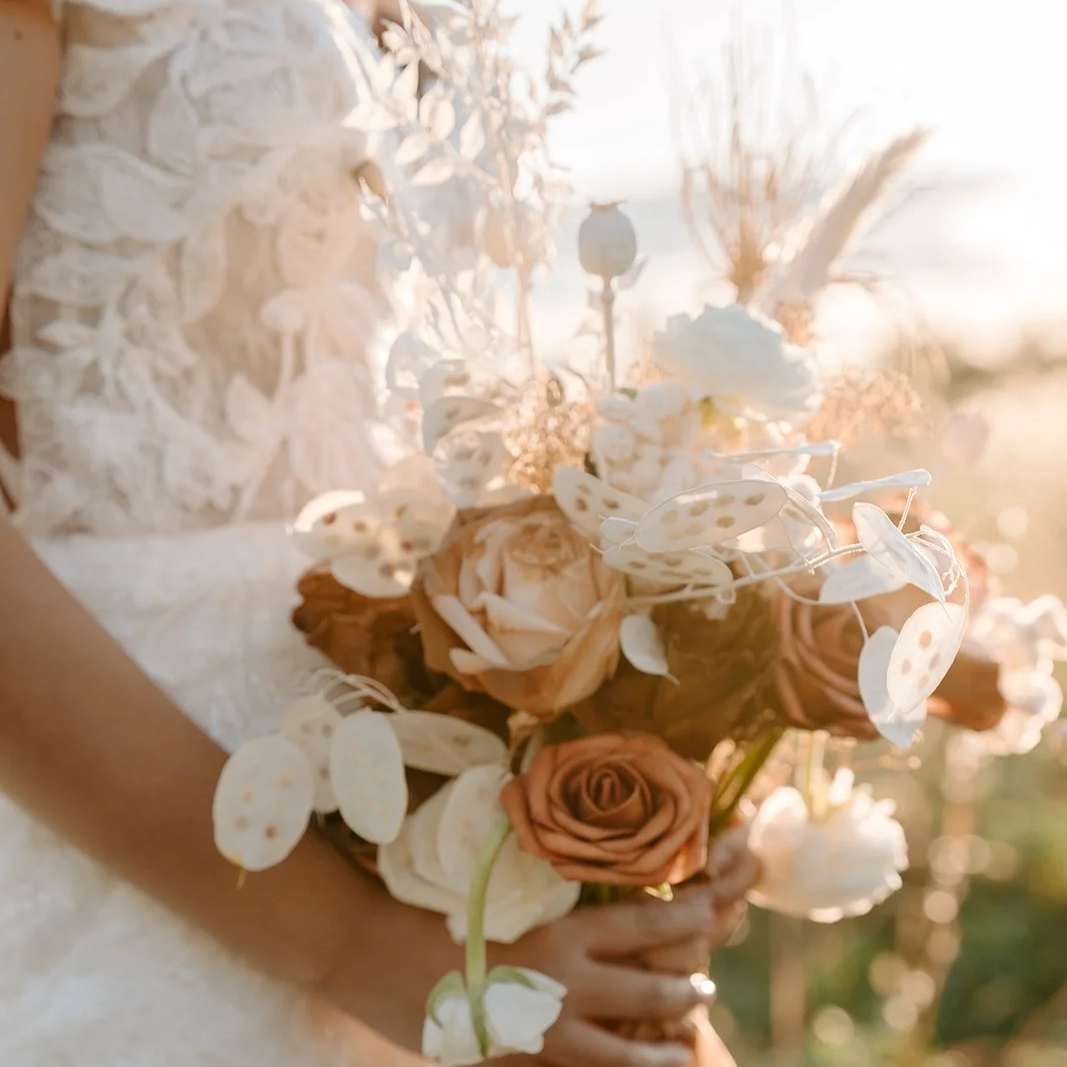 A person holding a bouquet of flowers, including roses and other blooms, with a blurred outdoor background and sunlight illuminating the scene.