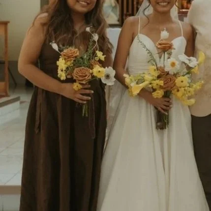 Two women holding bouquets of yellow and brown flowers, one in a dark dress and the other in a white dress, at a social event or celebration.