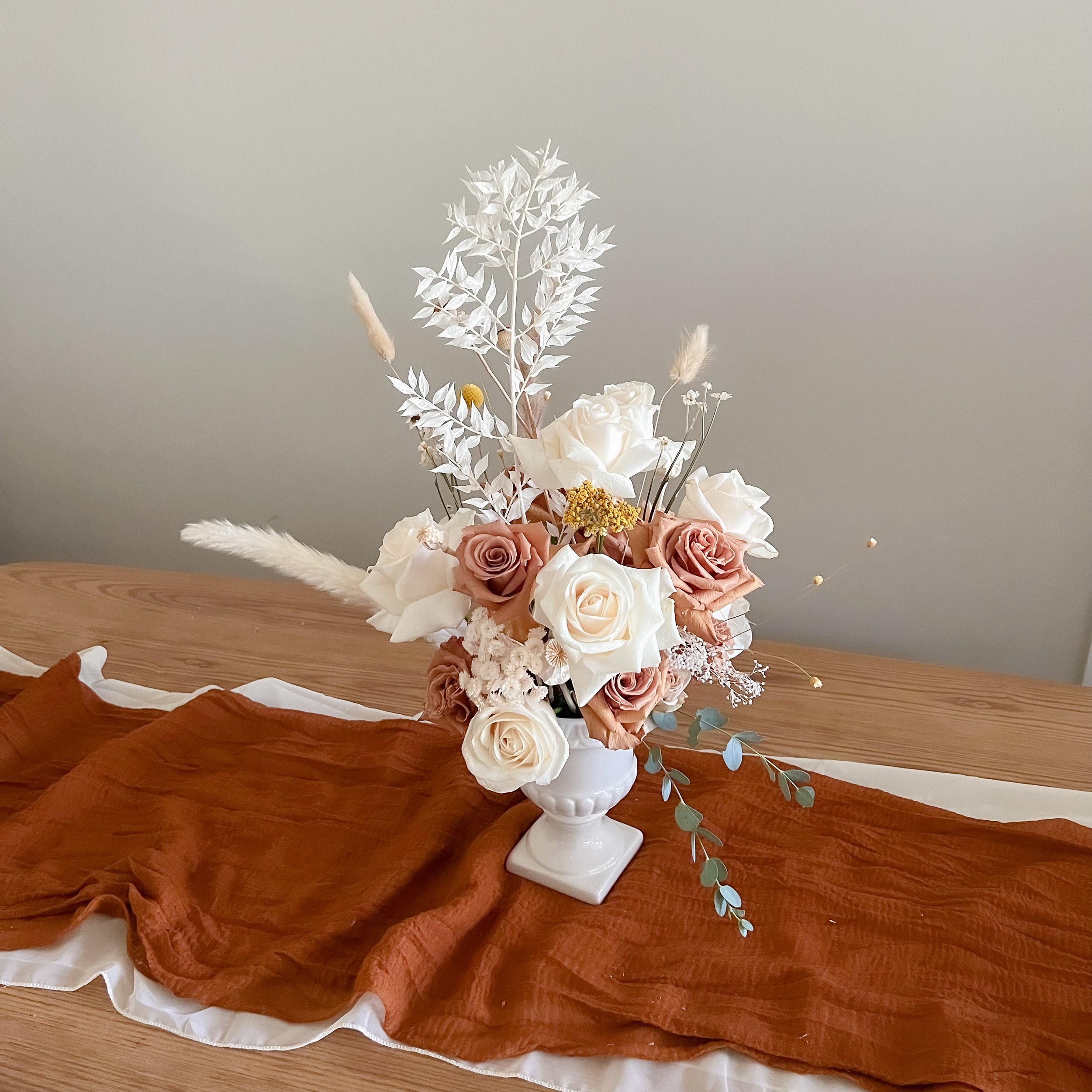 A floral arrangement in a white vase on a wooden surface, featuring roses, dried grasses, and white foliage, with a draped brown fabric underneath.