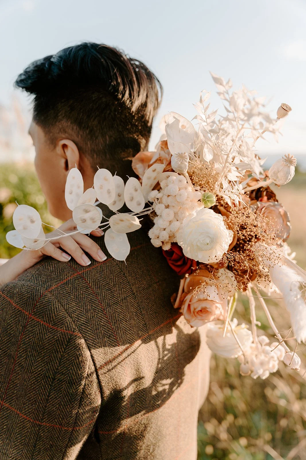 A close-up of a woman holding a large bouquet of white and blush pink flowers while embracing a man, outdoors during sunset.