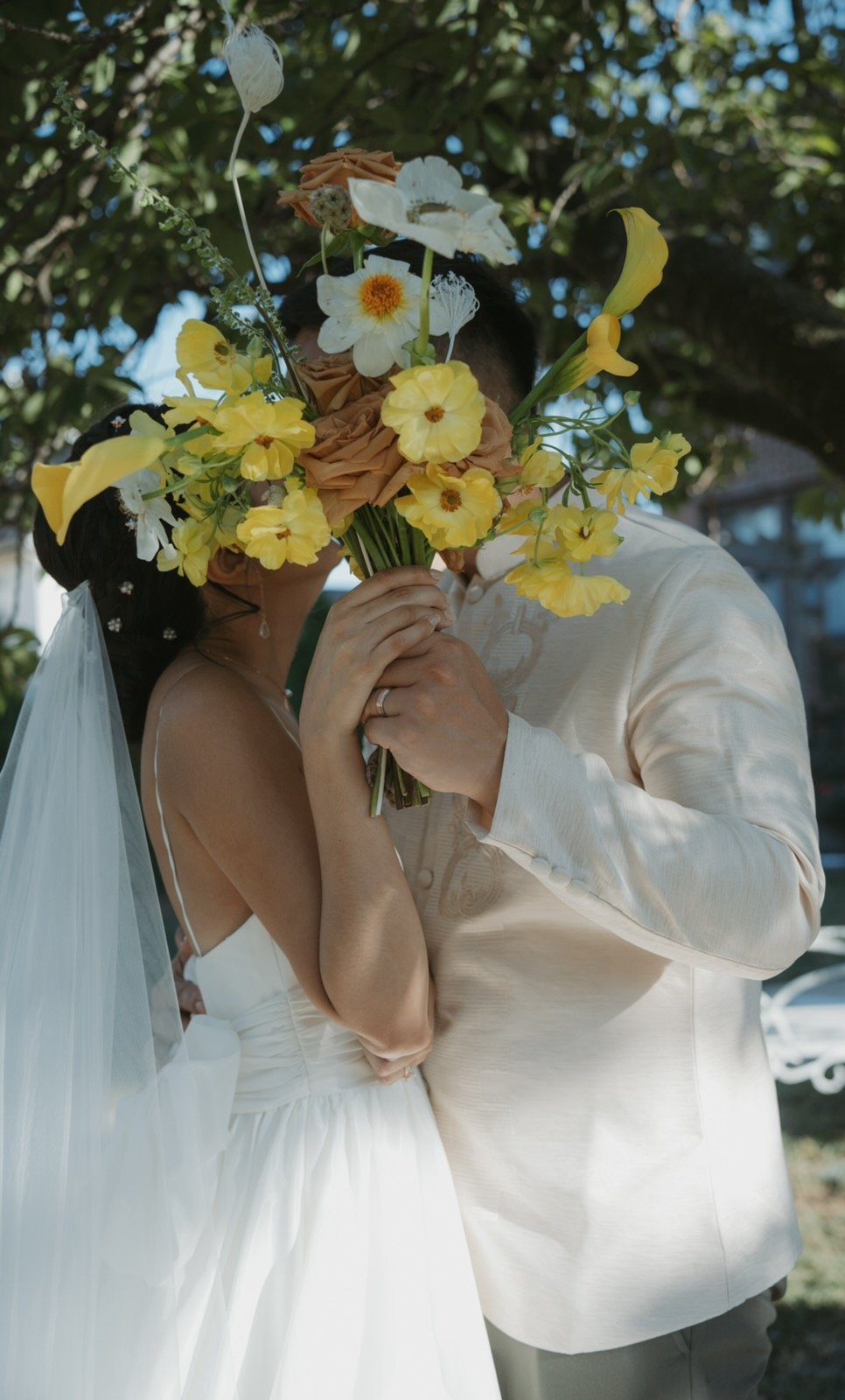 A couple in wedding attire holding a large bouquet of flowers in front of their faces during an outdoor ceremony.
