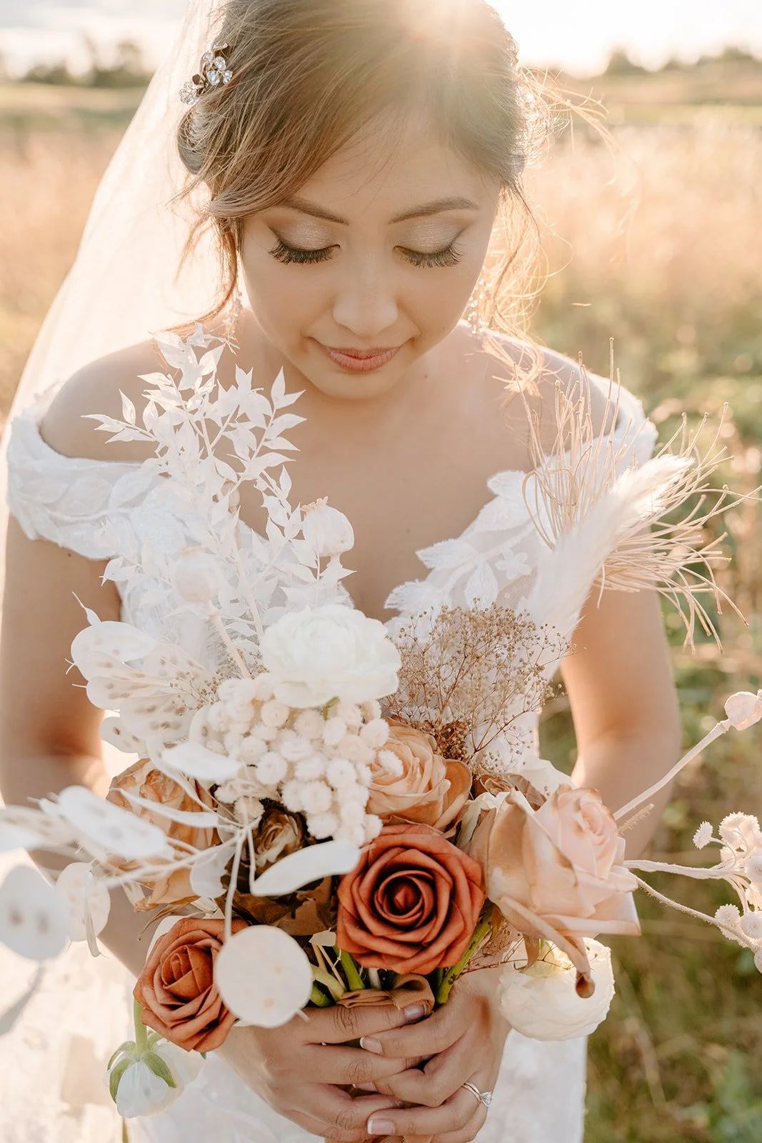 A bride holding a bouquet of peach, white, and cream flowers with greenery and dried elements, outdoors at sunset.