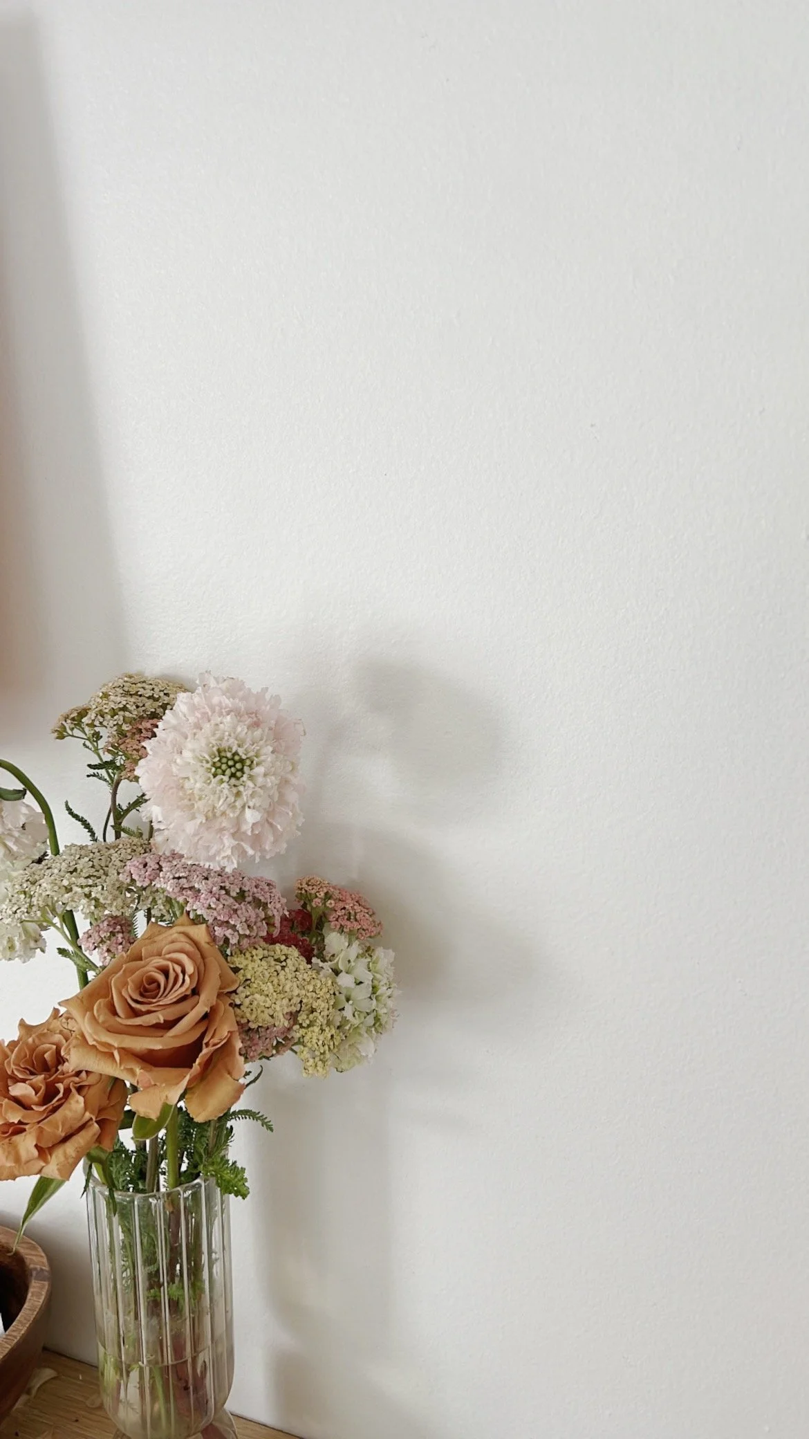 A bouquet of various flowers, including roses and hydrangeas, in a clear glass vase against a white wall.