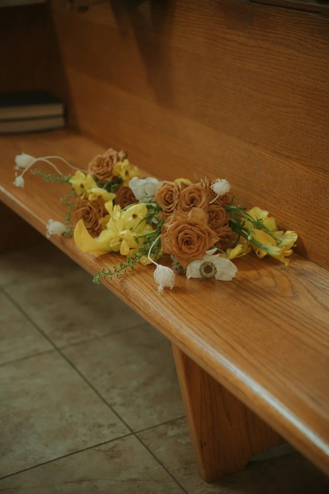 A floral arrangement of yellow, white, and brownish roses and other flowers rests on a wooden church pew.