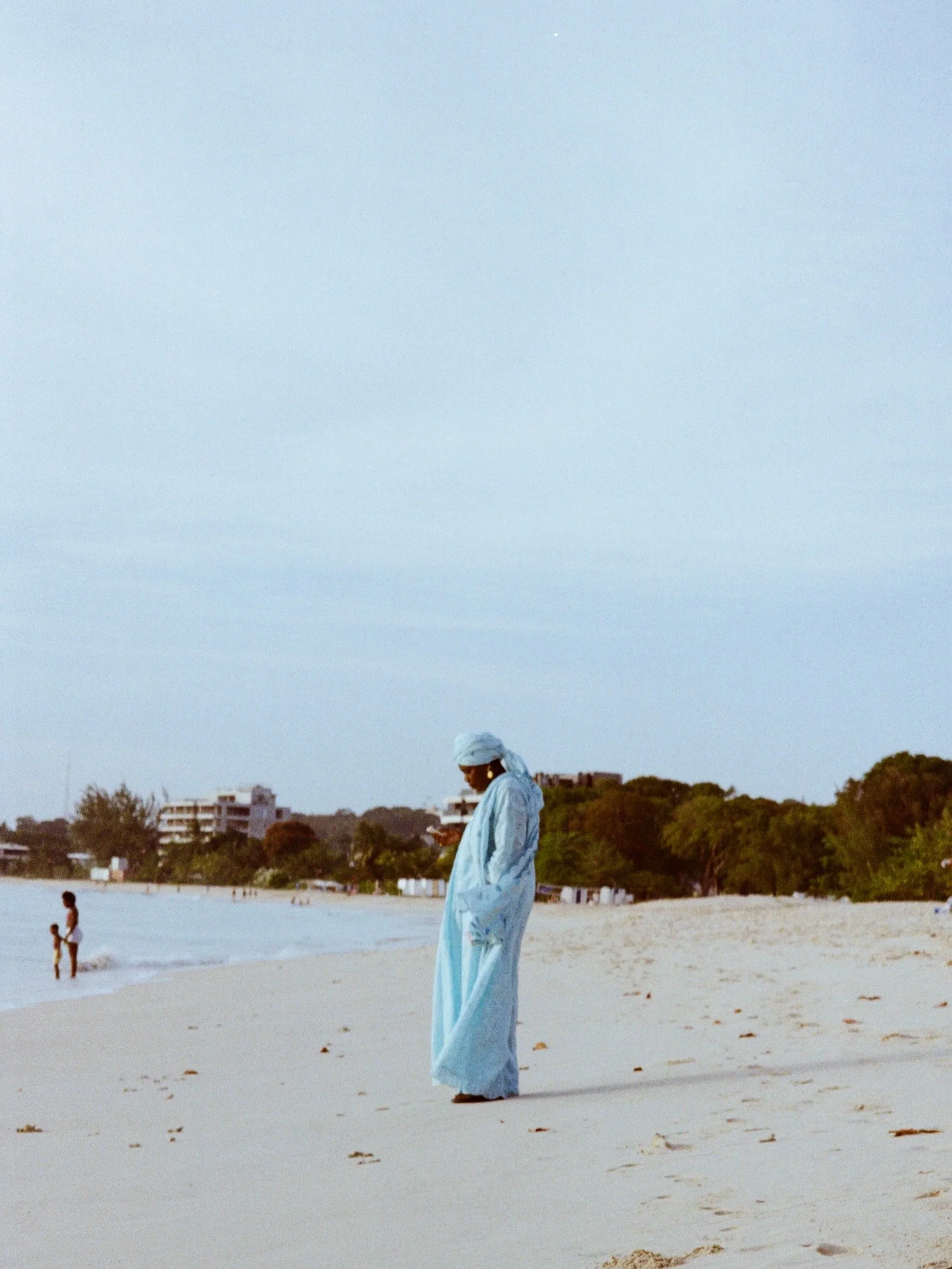 A woman in a light blue traditional dress and white headscarf standing on a sandy beach, with trees and buildings in the background, and a cloudy sky overhead.