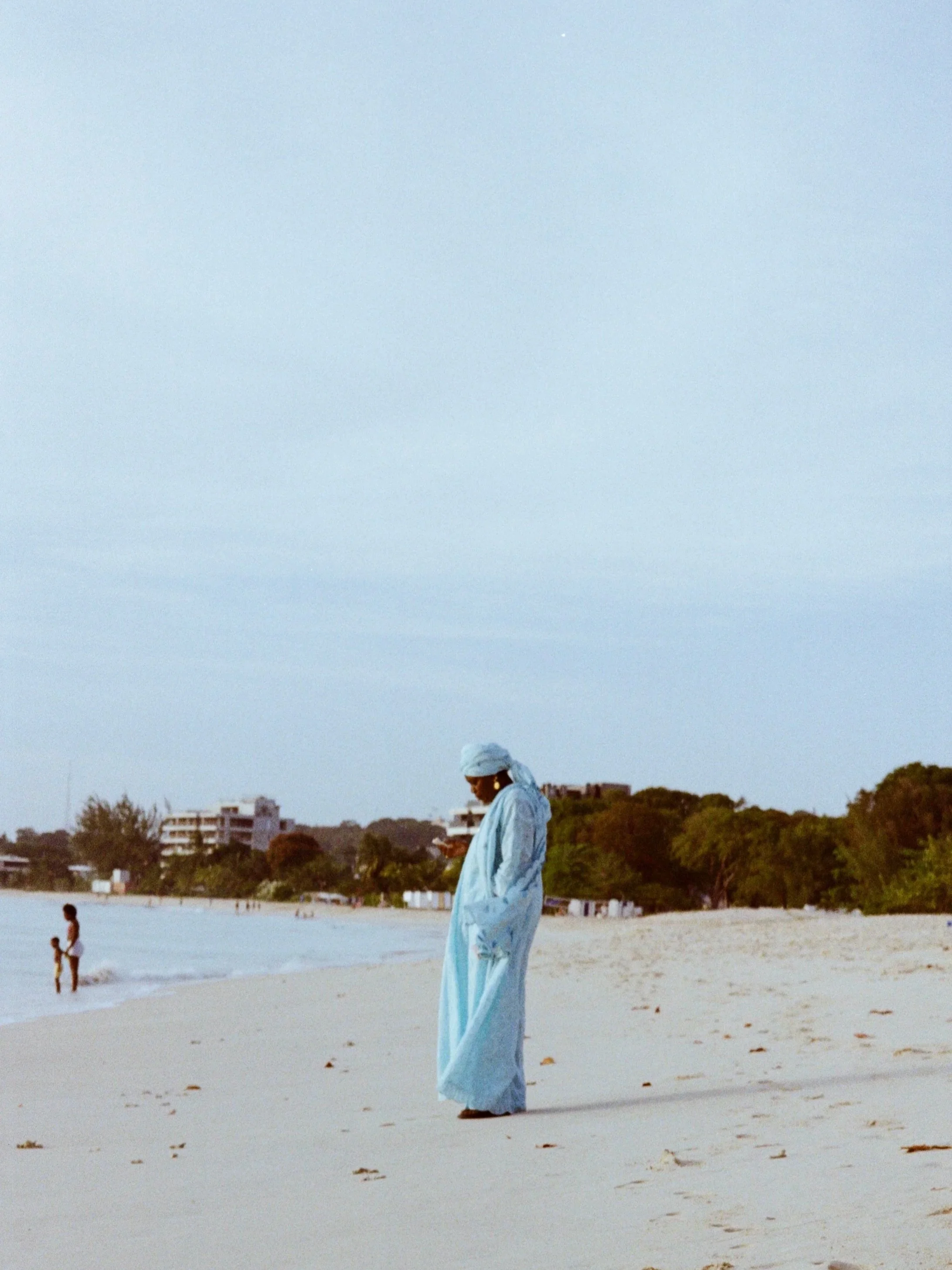 A woman wearing traditional light blue attire and head covering stands on a beach, with her head bowed and hands in her pockets, in front of a calm ocean scene with children playing nearby, trees, and buildings in the background during daytime.