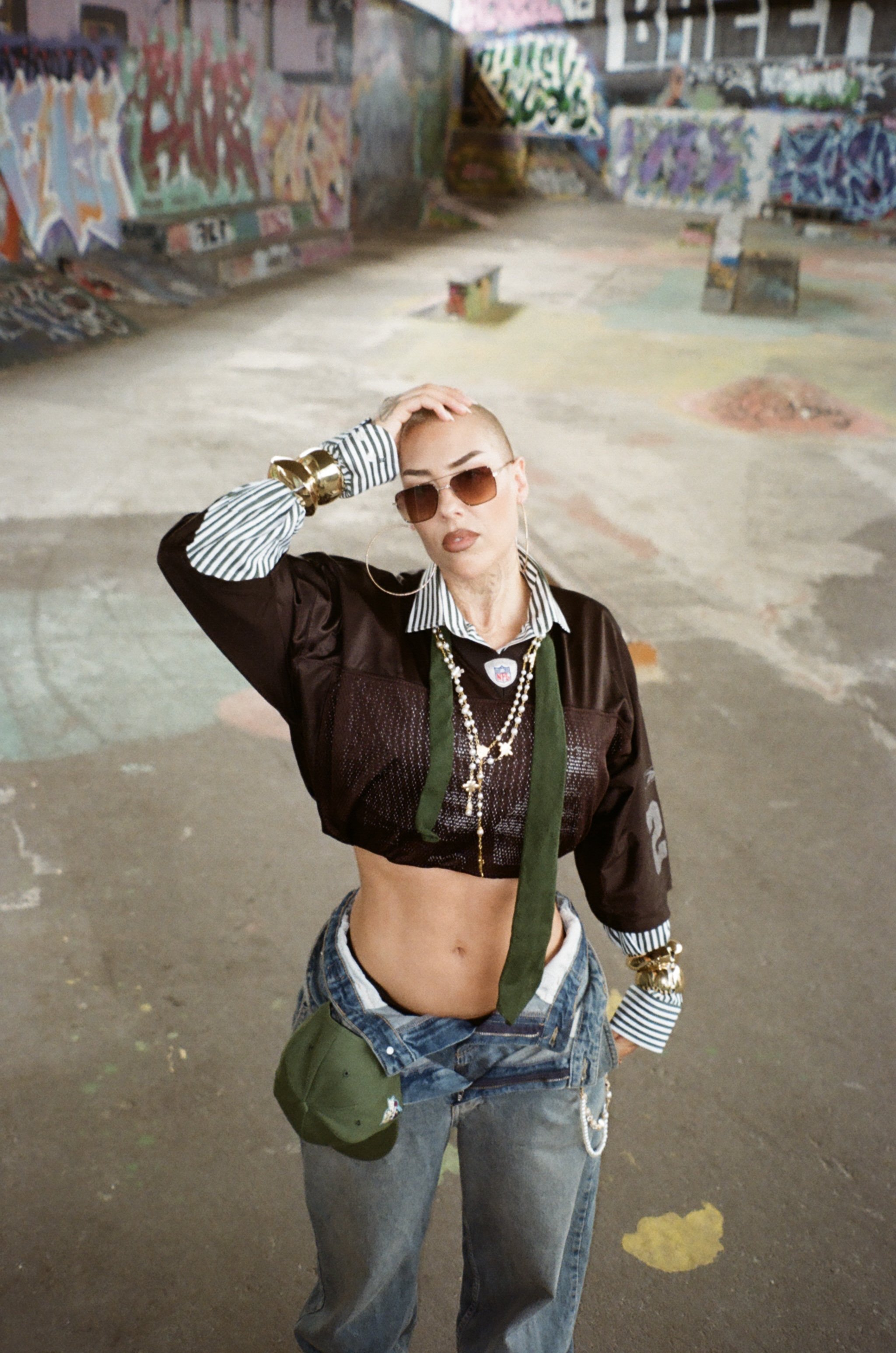 A young woman with a shaved head wearing sunglasses, a cropped dark jacket with stripes on the sleeves, gold jewelry, a crop top revealing her midriff, and loose jeans with a chain wallet, standing in an art-filled skate park with colorful graffiti on the walls and skate ramps in the background.