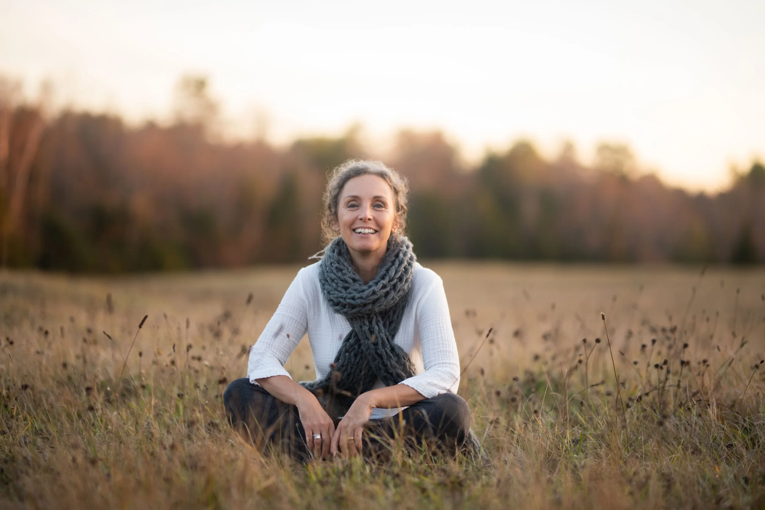 A woman smiling while sitting cross-legged in a field, wearing a white top and gray scarf, with trees in the background at sunset.