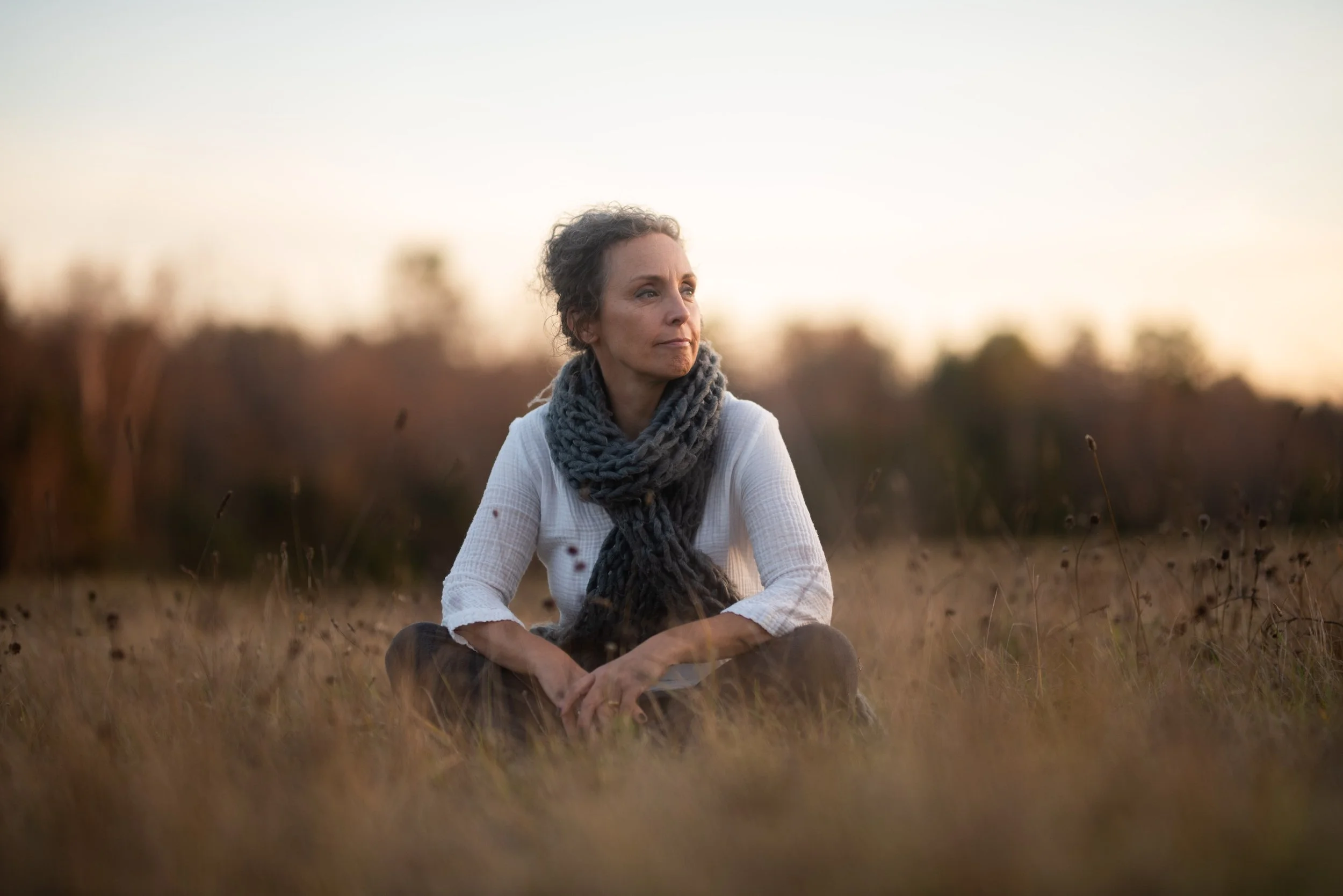 A woman sitting cross-legged in a field during sunset, wearing a white top and a chunky knitted scarf, looking thoughtfully into the distance with trees blurred in the background.