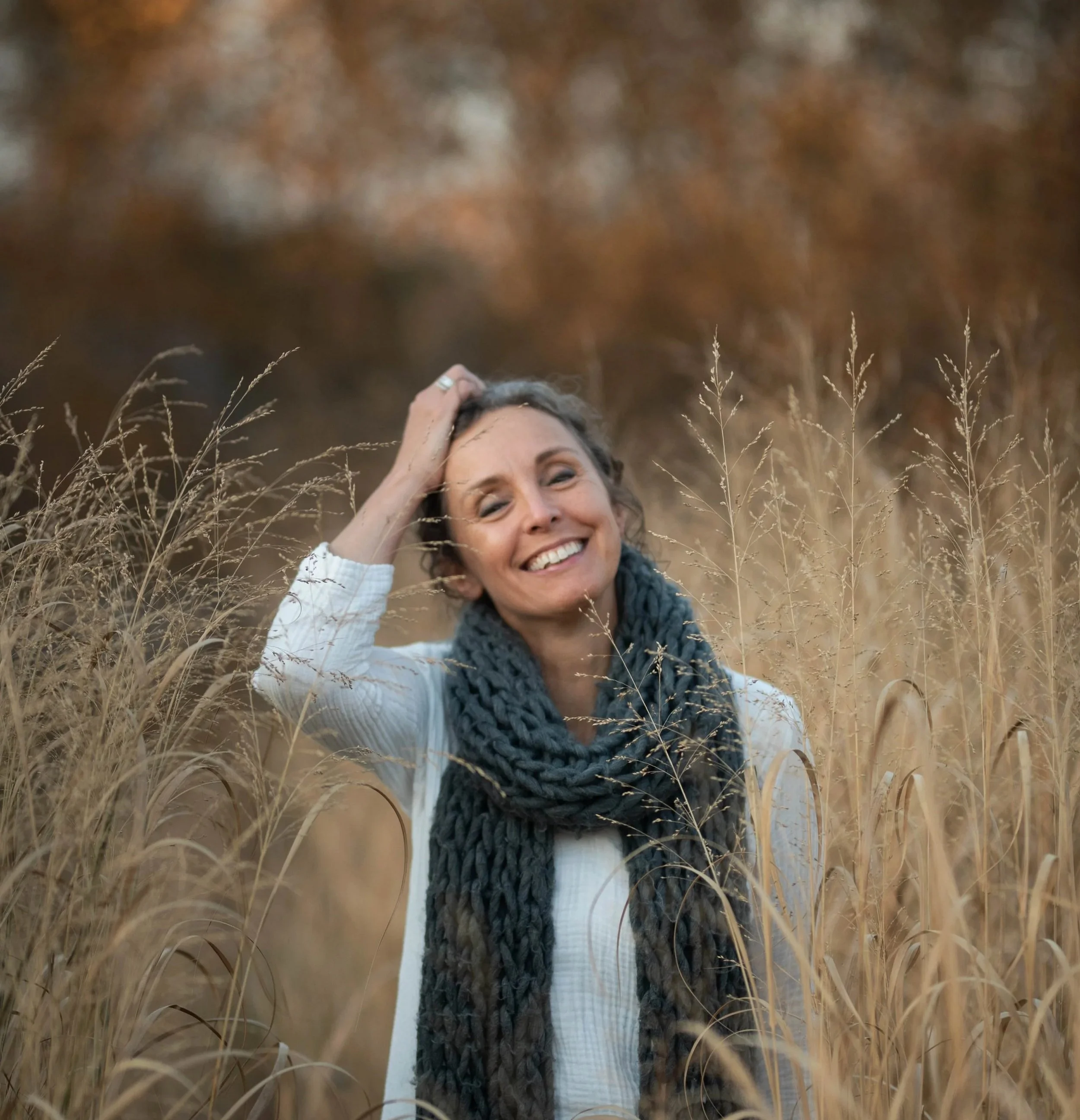 A woman with curly hair smiling, wearing a white top and a dark knitted scarf, standing in a field of tall, dry grass with a blurred autumn-colored background.