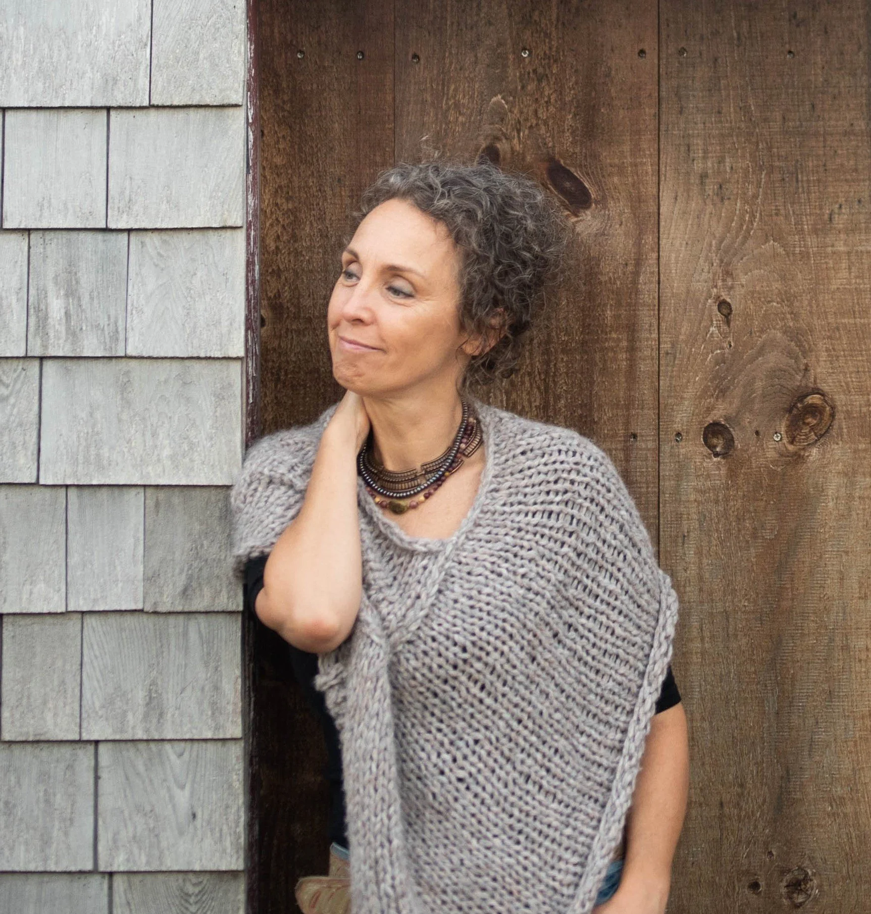 A woman with short, curly gray hair, wearing a chunky knit sweater, layered necklaces, and a bracelet, standing in front of a background of wooden and shingle siding, looking off to the side with a gentle smile.