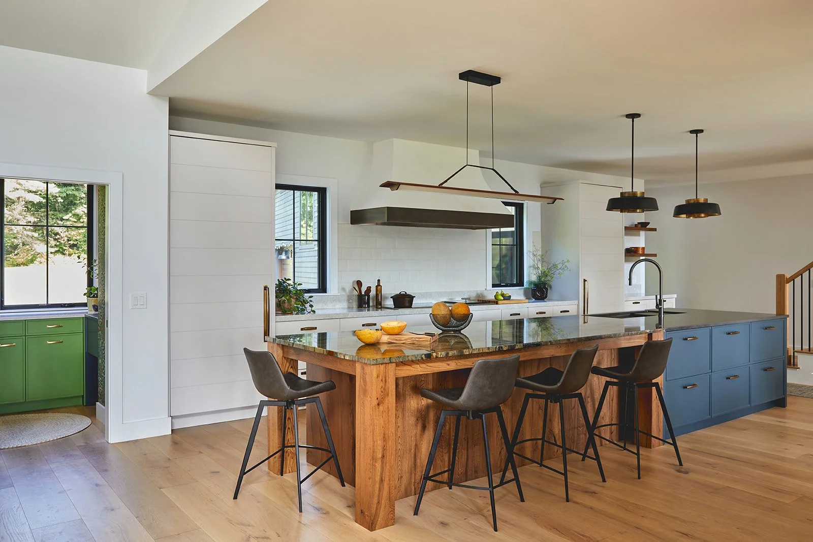 Modern kitchen with a wooden island, black bar stools, and white cabinetry. Green and blue accents, black pendant lights, and natural light from windows.