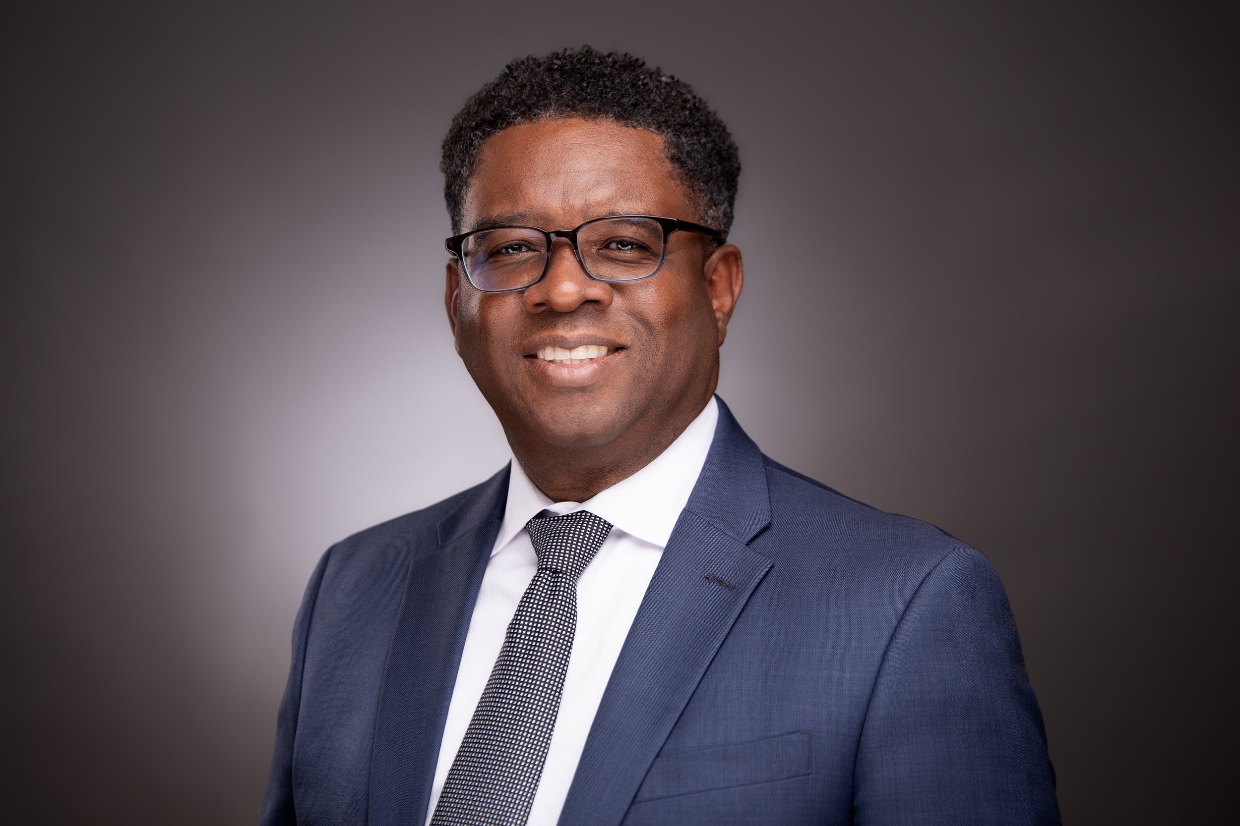 A professional man with dark curly hair, wearing glasses, a navy blue suit, white shirt, and patterned tie, smiling against a plain dark background.