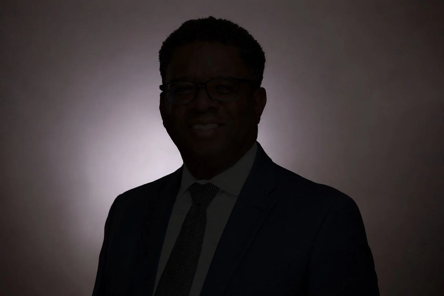 A dark silhouette of a man wearing glasses, a suit, and a tie, standing in front of a light background.