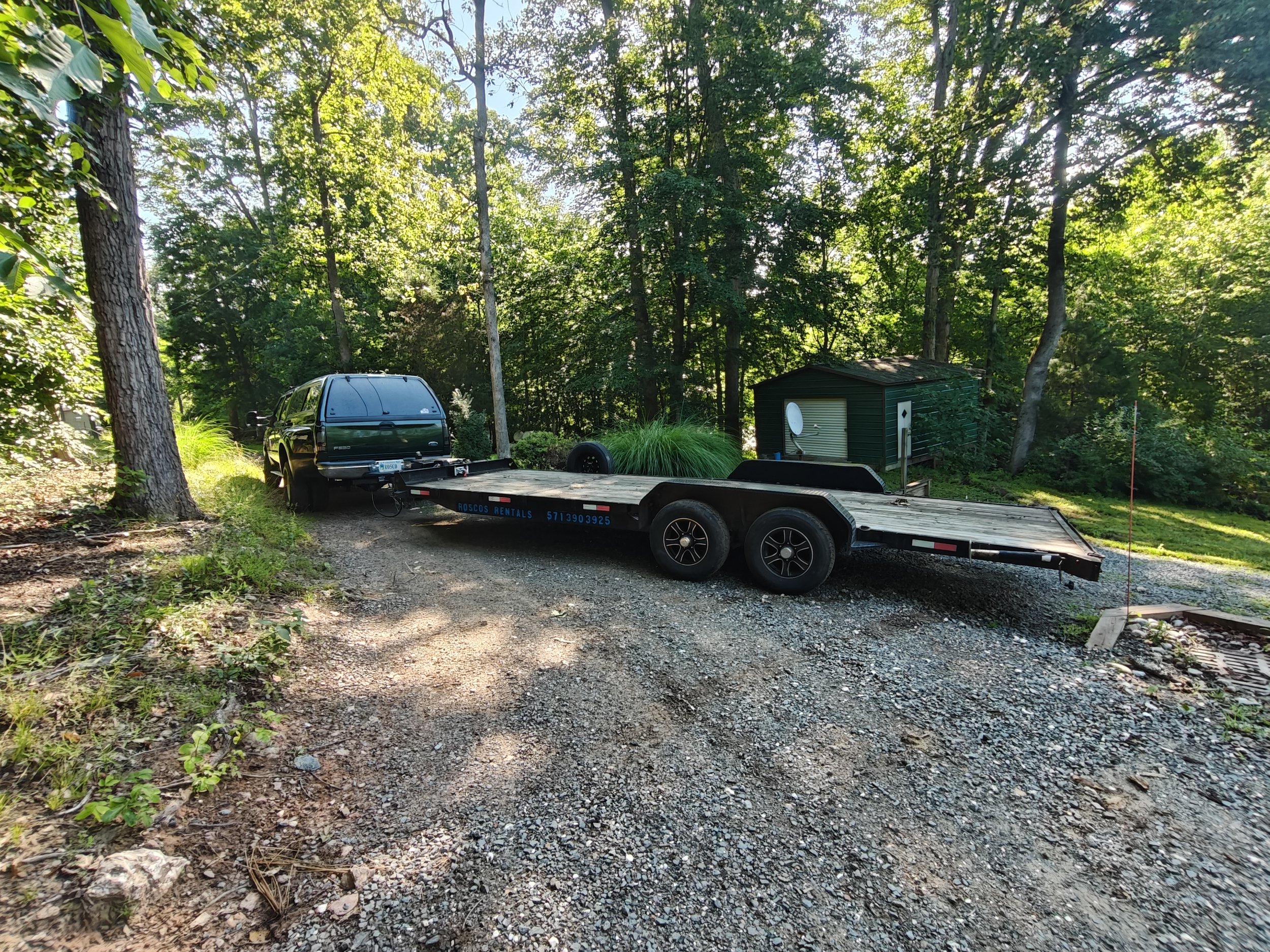 A black truck with a flatbed trailer parked on a dirt and gravel driveway surrounded by green trees and foliage. A small dark-colored shed with a white satellite dish on its side is visible in the background.