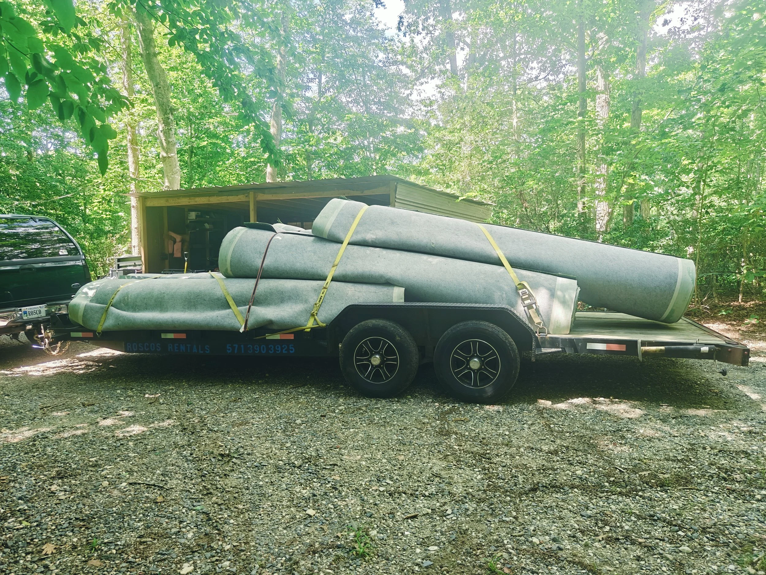 A trailer carrying large rolled-up carpet and other materials is parked on a gravel surface in a wooded area with tall trees and green foliage.