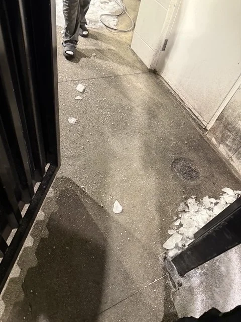 Concrete sidewalk with debris and ice near an open gate and a wall, with part of a person's legs visible.