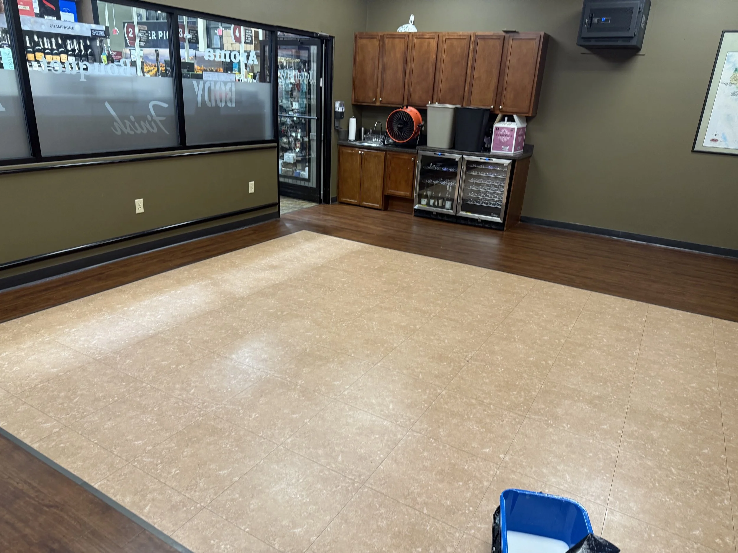 Empty restaurant or cafe interior with brown and beige flooring, a small kitchenette with wooden cabinets, a sink, a paper towel roll, a fan, and beverage coolers. Part of a blue chair is visible in the bottom right corner.