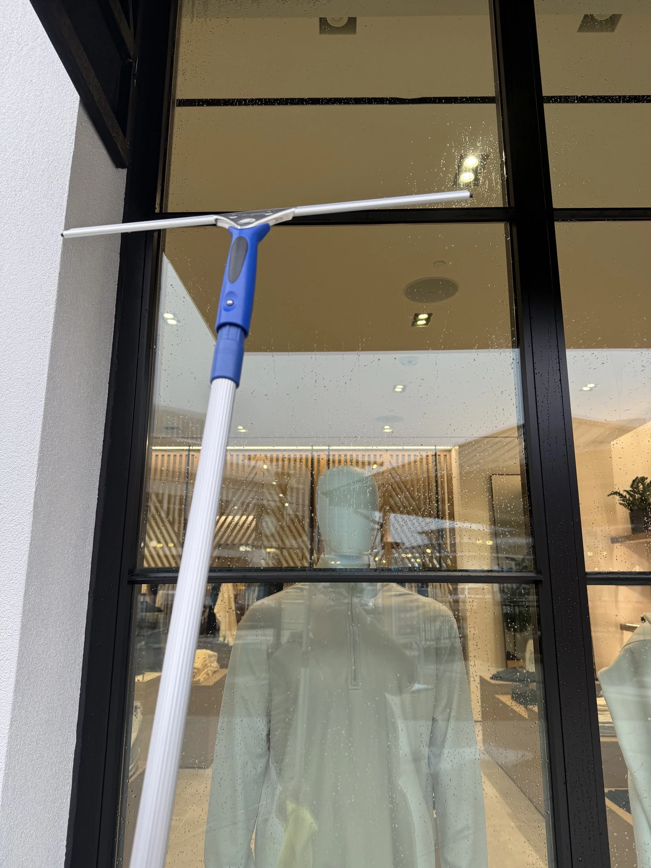 A mannequin is seen through a large glass window with raindrops on it. The mannequin is inside a building, and the reflection of ceiling lights is visible on the window. The person is wearing a shirt and is facing forward.