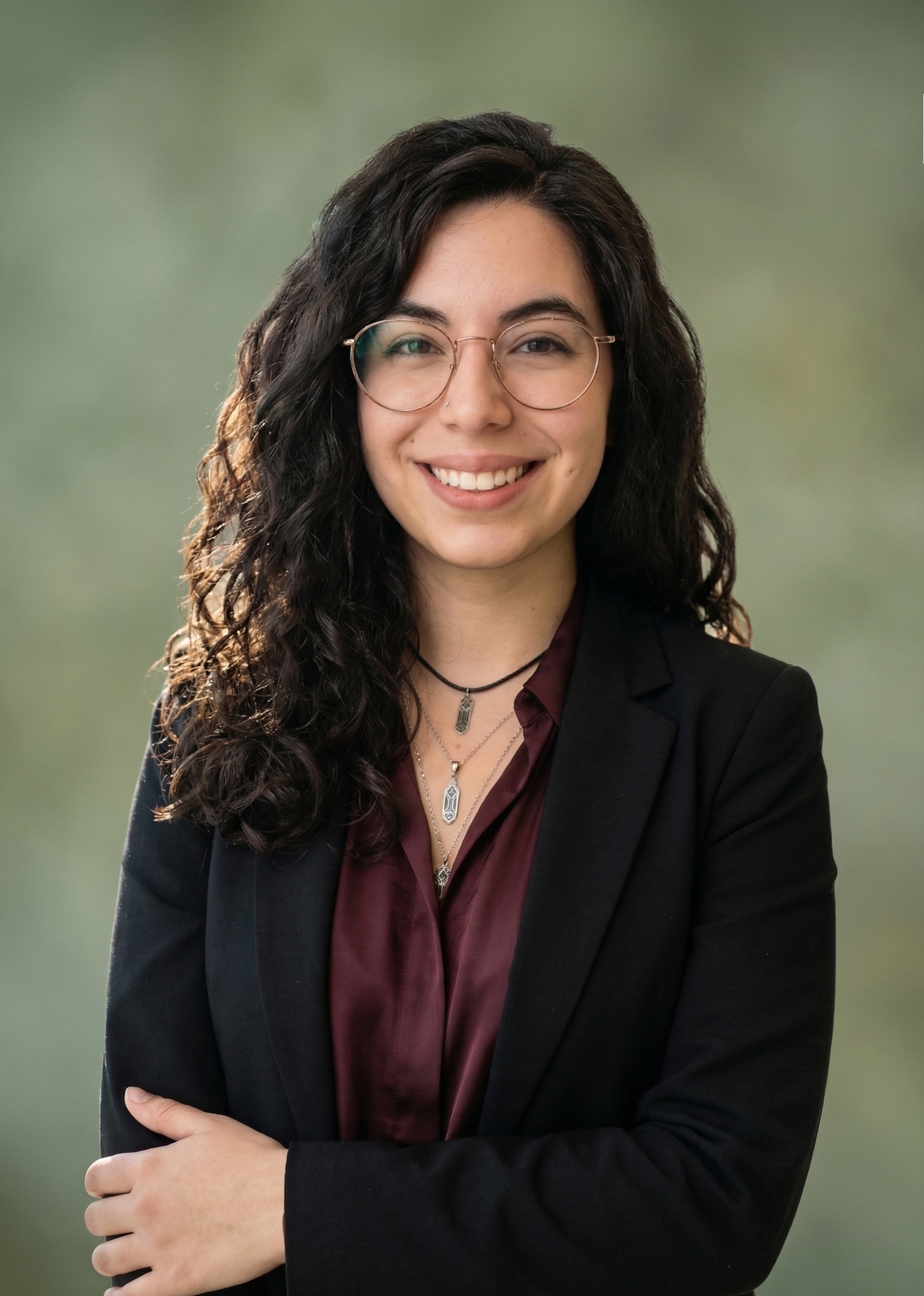 A woman with dark curly hair and glasses, wearing a dark blazer over a maroon shirt, smiling and crossing her arms, standing outdoors with a blurred green background.