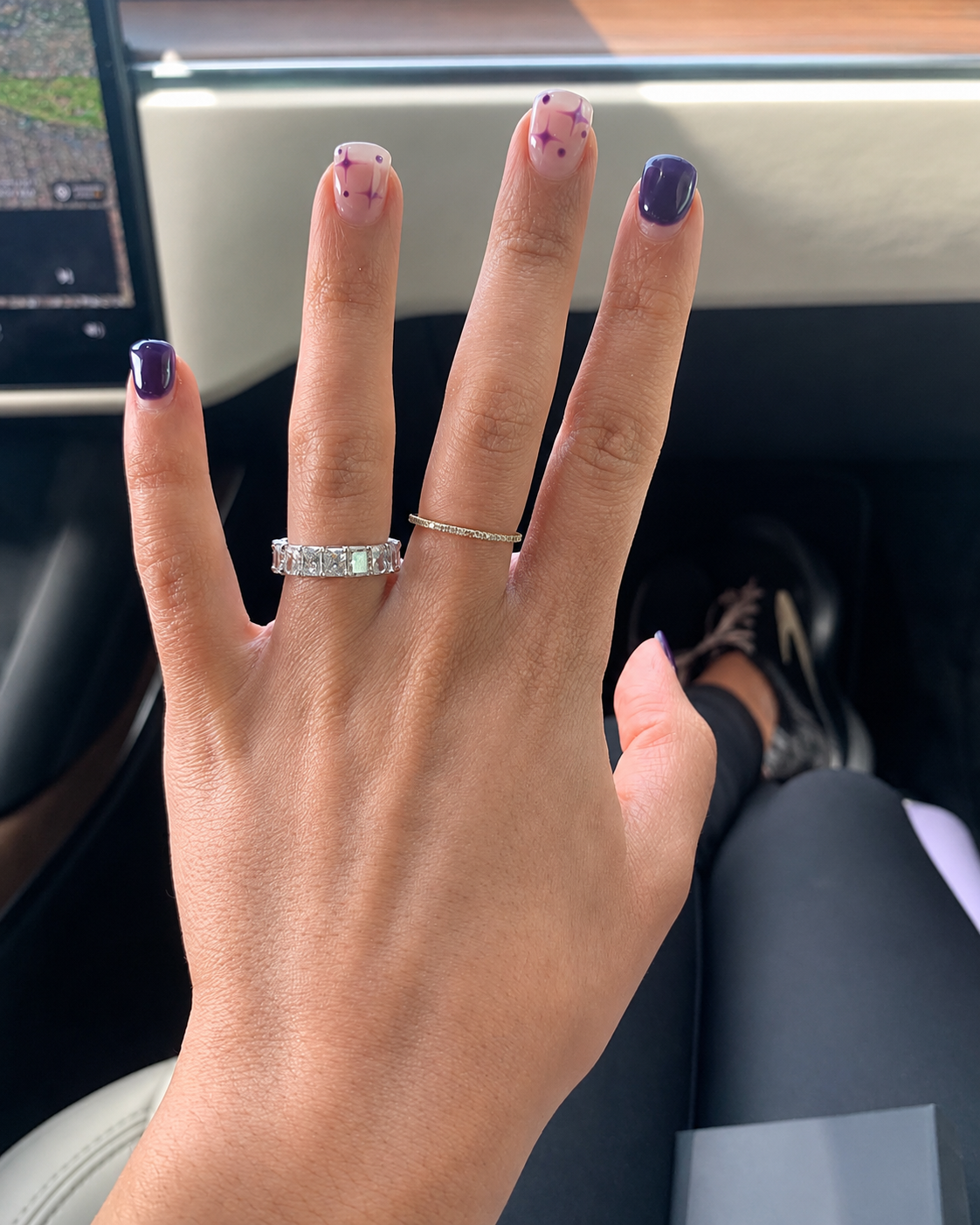 A person's left hand showing three rings and nail art with purple and pink colors. The person is inside a vehicle, with a section of a computer screen visible in the background.