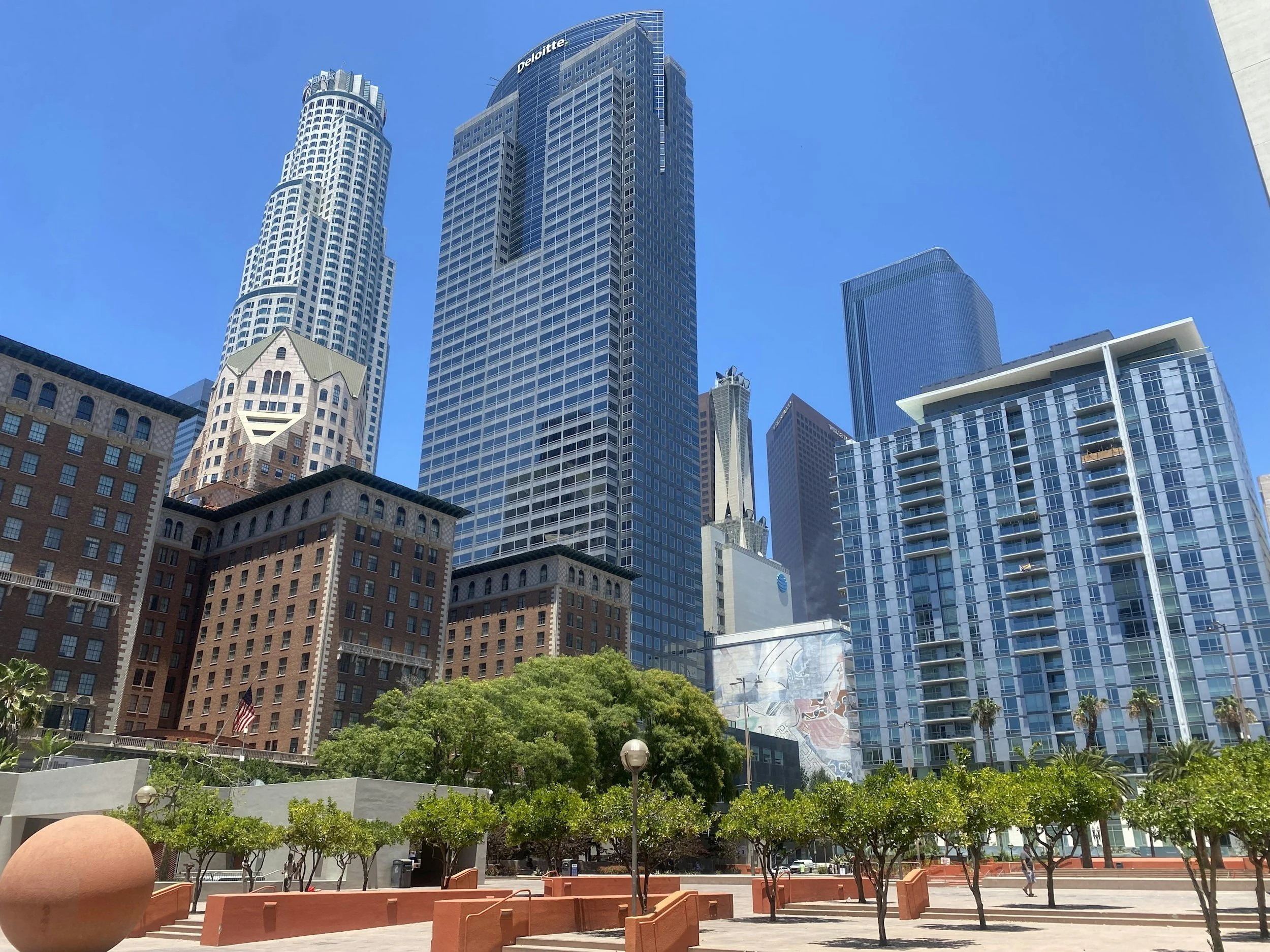 A cityscape with tall skyscrapers and modern buildings under a clear blue sky, with some greenery and a small park area in the foreground.