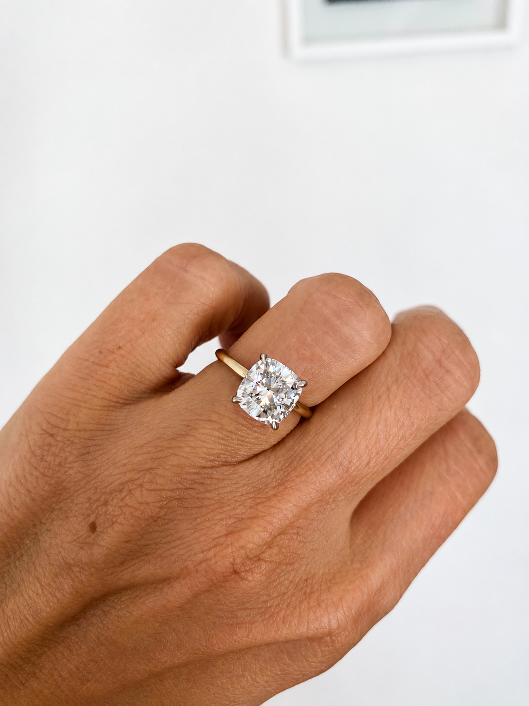 Close-up of a hand wearing a gold ring with a large, square cut diamond solitaire on a plain, light background.