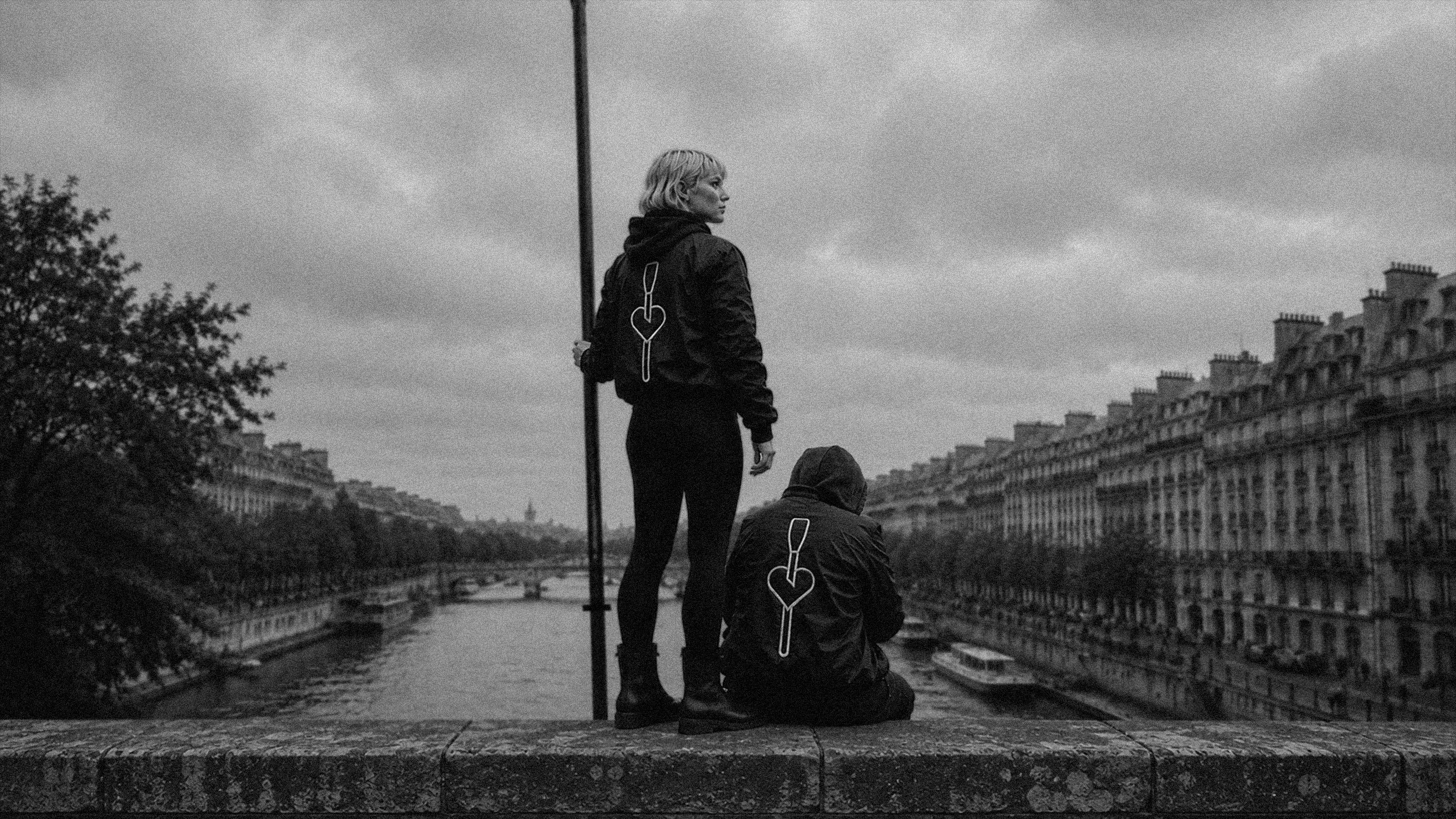 Deux personnes sur un pont au bord de l'eau, l'une debout et l'autre assise, avec des bâtiments anciens en arrière-plan et un ciel nuageux, en noir et blanc.