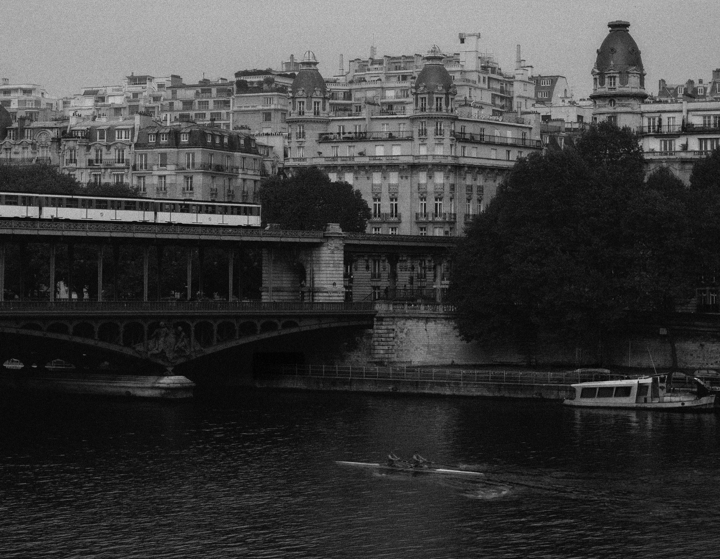 Vue d'une ville avec des bâtiments haussmanniens et un train traversant un pont au bord de la Seine, en noir et blanc.