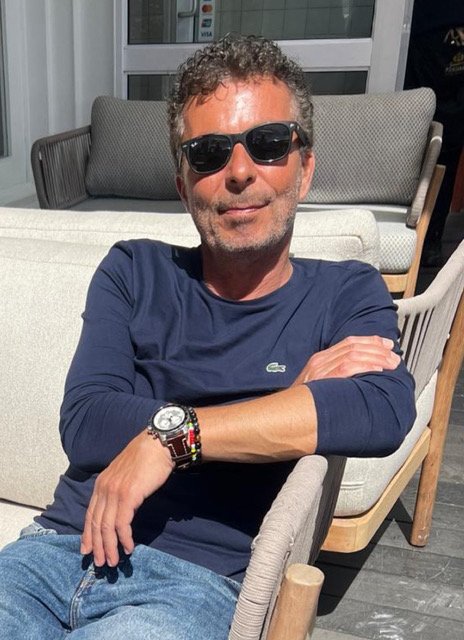 Man with curly hair and sunglasses sitting outdoors, crossing his arms, wearing a navy blue shirt and a watch, sitting on a wooden chair.