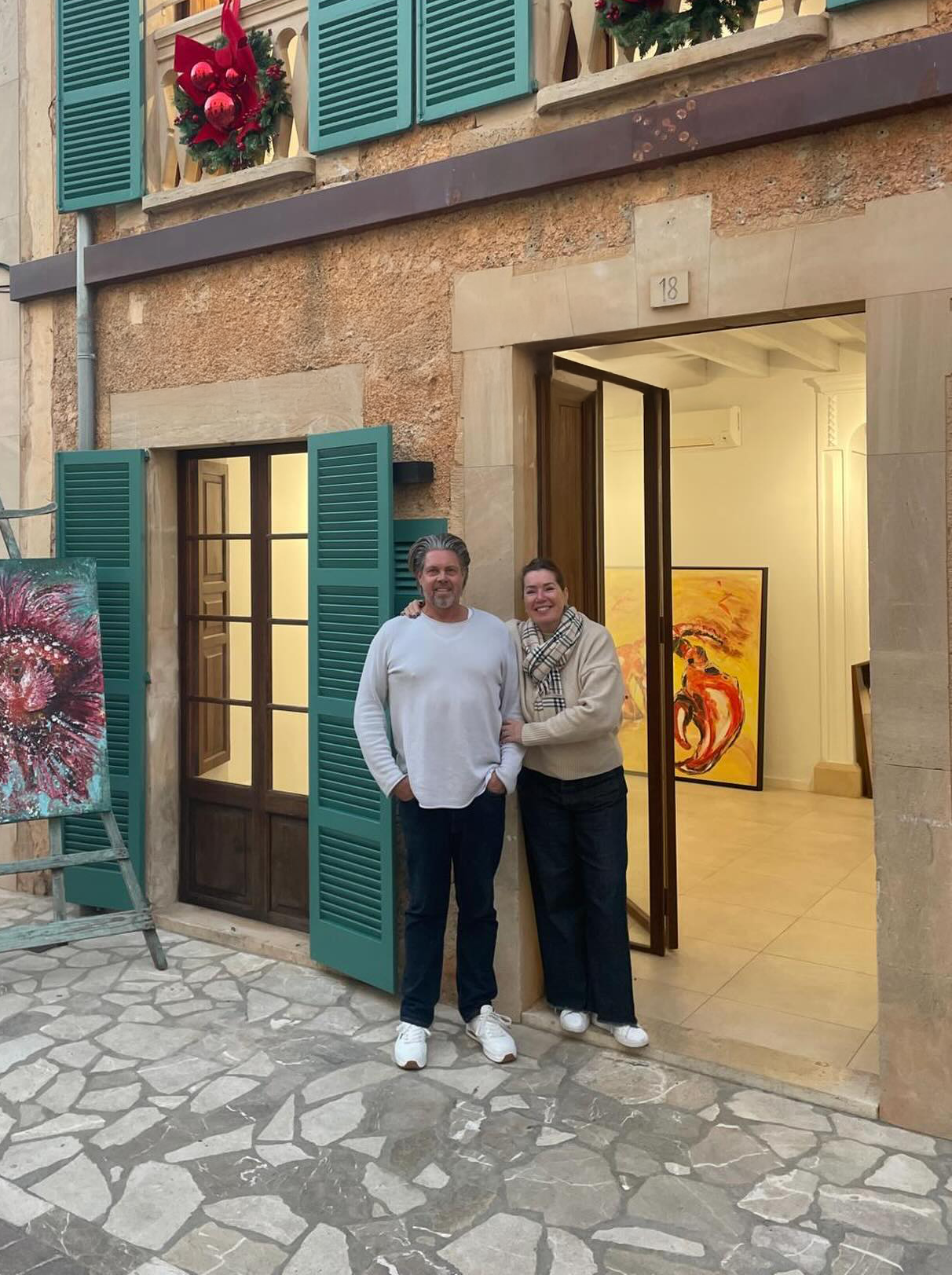 Two people stand smiling outside an art gallery or studio entrance, with paintings visible inside. The man is wearing a white sweater and jeans, and the woman is wearing a beige sweater, plaid scarf, and black pants. The building has a rustic appearance with turquoise shutters and Christmas decorations.