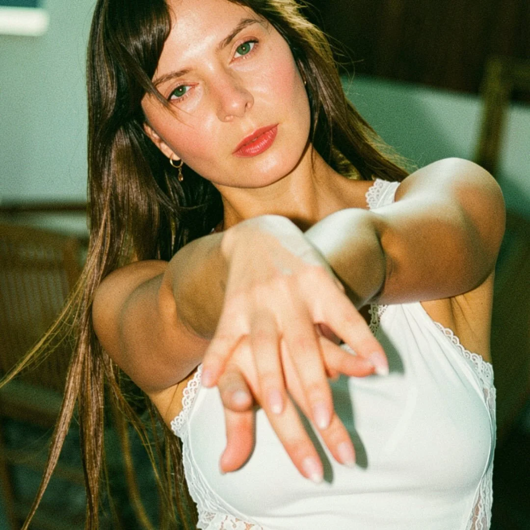 A young woman with brown hair and green eyes wearing a white sleeveless top, looking directly at the camera with her arms extended forward.