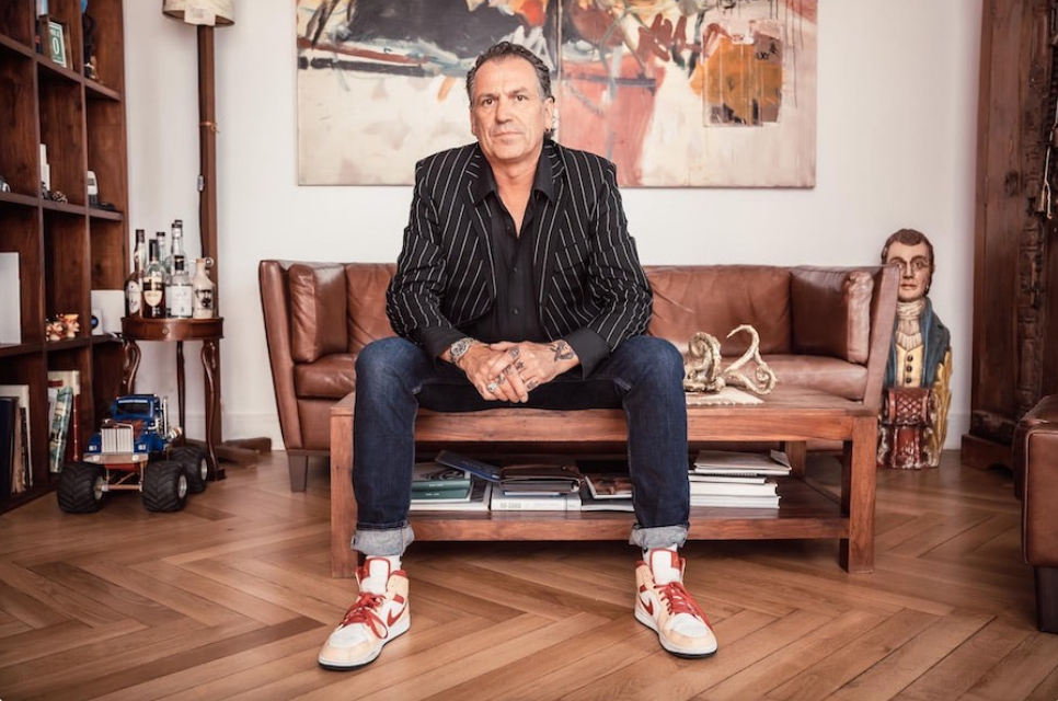 A man with tattoos on his hands sitting on a wooden coffee table in a living room. He is wearing a black pinstripe blazer, black shirt, blue jeans, and white and red sneakers. The room features a brown leather sofa, a painting hanging on the wall, decorative sculptures, books, and a large sculpture of a person's head in the background.