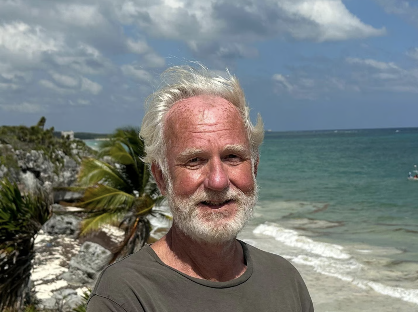 A smiling older man with white hair and beard standing on a tropical beach with ocean, palm trees, and cloudy sky in the background.