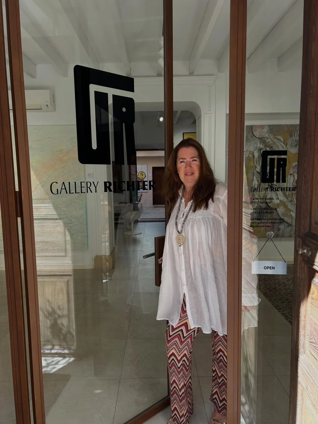 Woman standing inside an art gallery, visible through a glass door with the gallery's logo and contact information, wearing a white blouse and patterned pants.