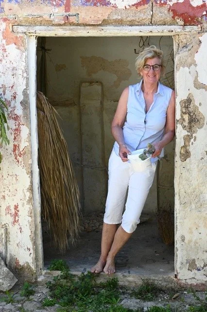 An older woman with glasses and short, curly gray hair standing barefoot in a doorway of an old, worn building with peeling paint. She is smiling and holding gardening tools, dressed in a light blue sleeveless shirt and white capris.