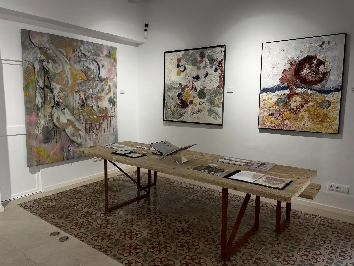Art gallery with three abstract paintings on the wall and a wooden table displaying art books and brochures.
