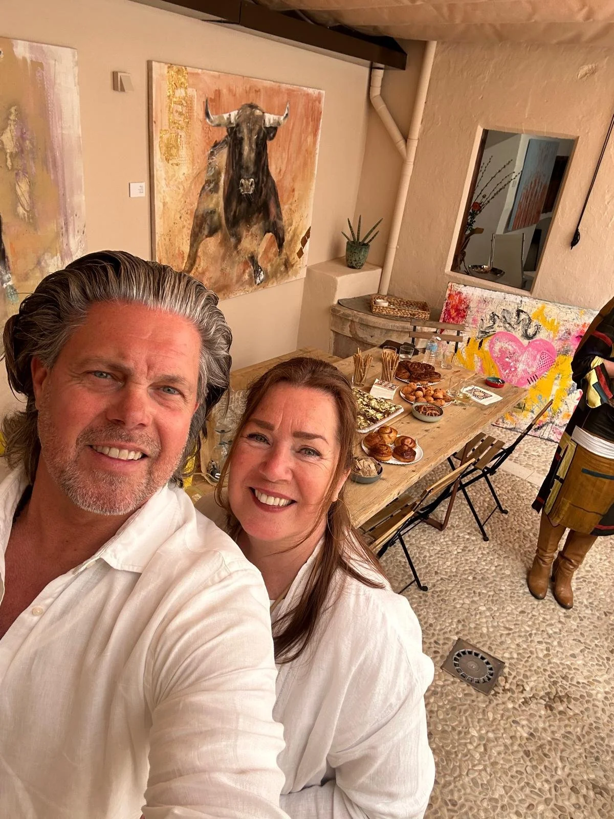 Two smiling people taking a selfie in a room with art and food on a table.
