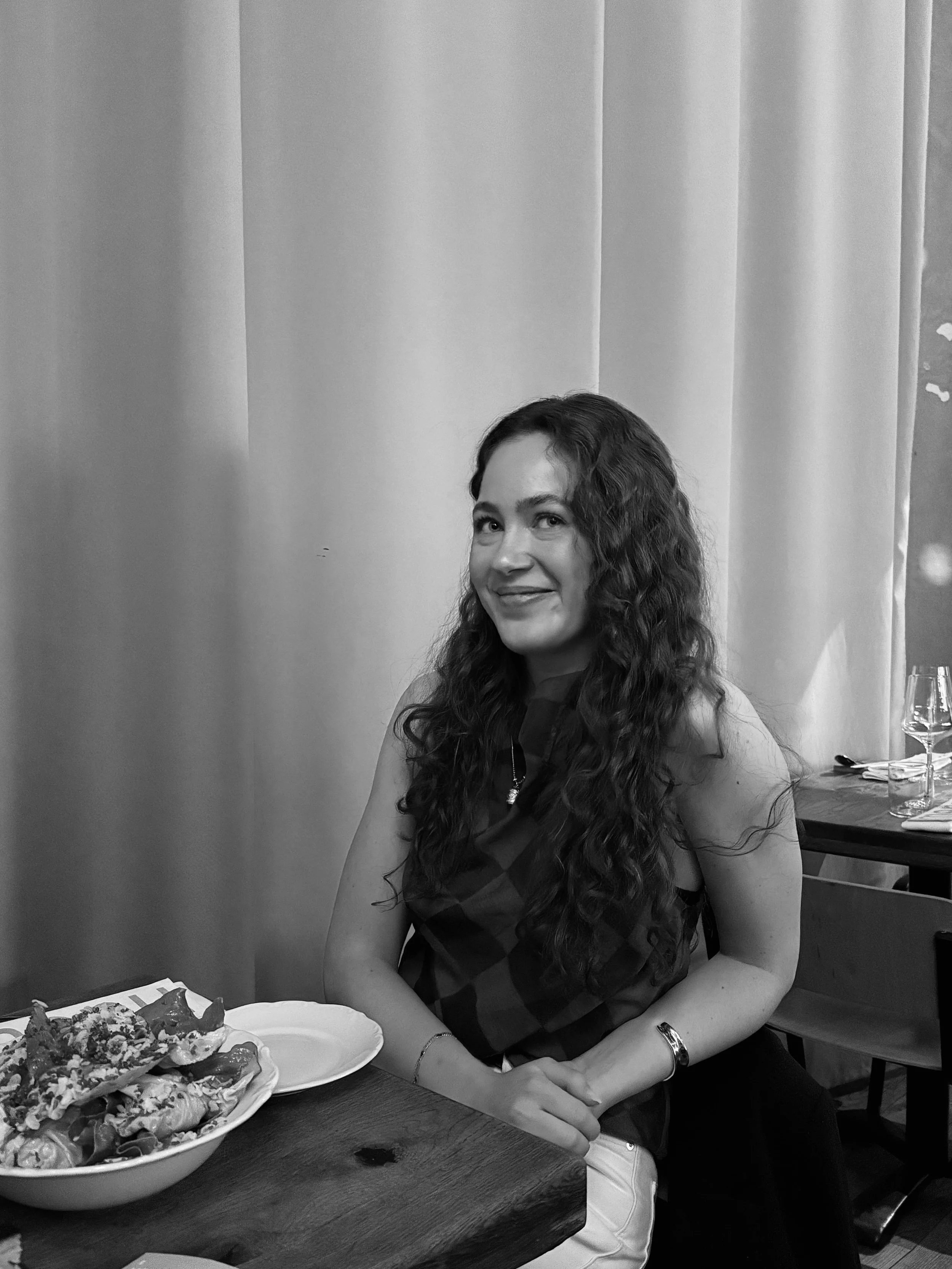 A woman with long curly hair sitting at a dining table in a restaurant. She is smiling and wearing a sleeveless top. On the table, there are plates of food, including pizza and salads, and wine glasses.