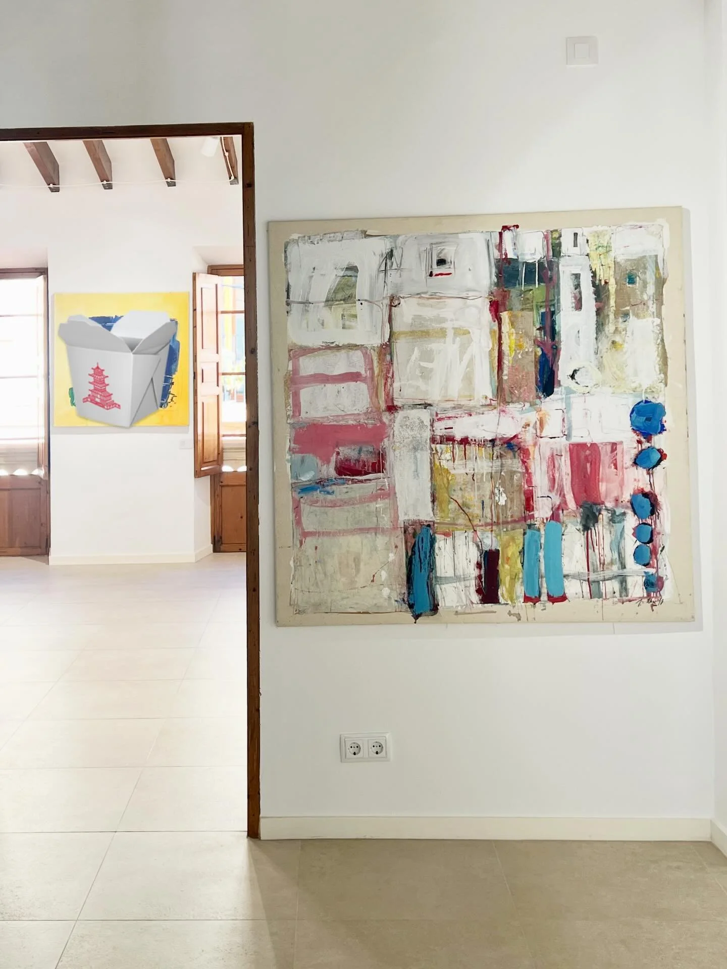 A gallery wall with an abstract painting featuring various colors including red, white, yellow, blue, and green, with a small painting of a white box with a red pagoda on a yellow background visible in the adjacent room.