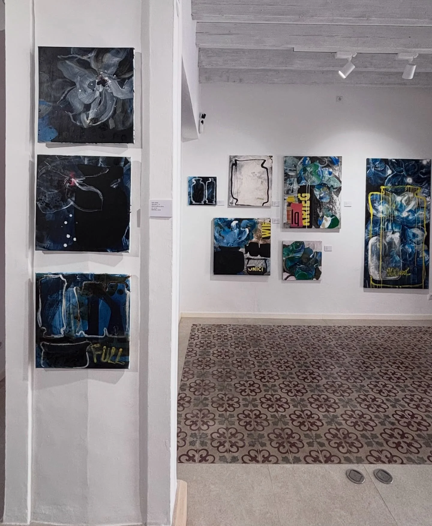 Gallery with abstract artwork on white walls, featuring bold blue, black, white, and green colors, and various patterns. The floor has patterned tiles in black, white, and gray.