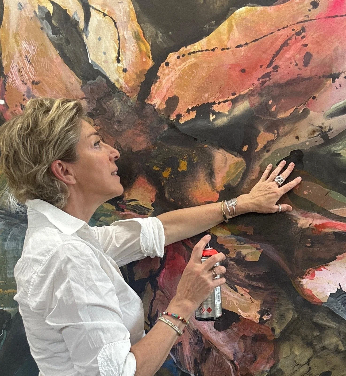 A woman with short blonde hair wearing a white shirt is spray painting an abstract, colorful artwork on a large canvas, using black spray paint and holding an aerosol can in her right hand.