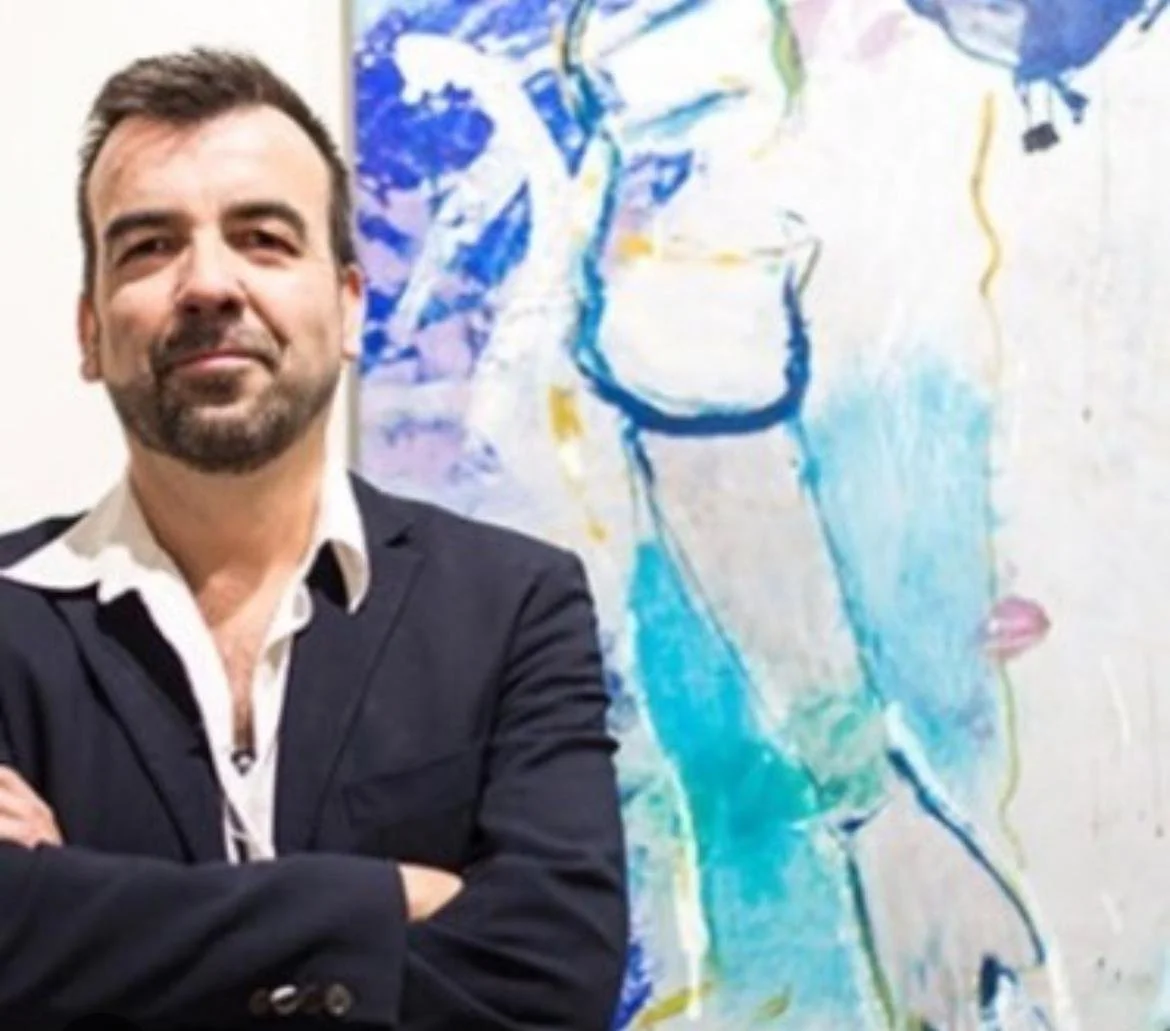 A man with dark hair and a beard, wearing a black blazer over a white shirt, standing with arms crossed in front of an abstract colorful painting of a person's face with blue, purple, yellow, and green hues.