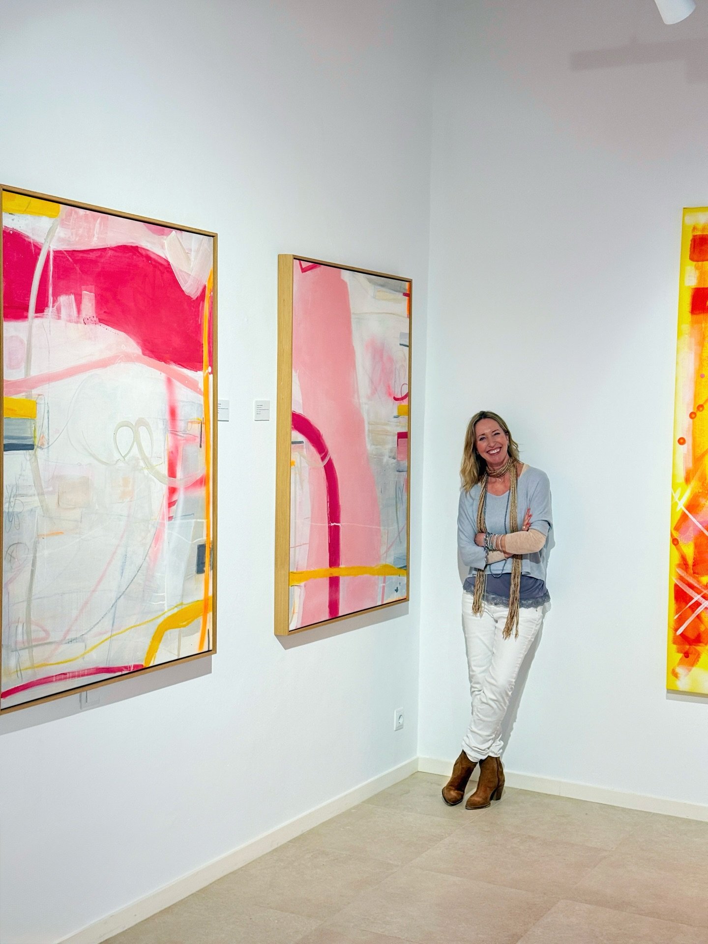 A woman standing in an art gallery with abstract colorful paintings on white walls, smiling with her arms crossed. She is wearing a light blue vest, white pants, brown boots, and accessories.